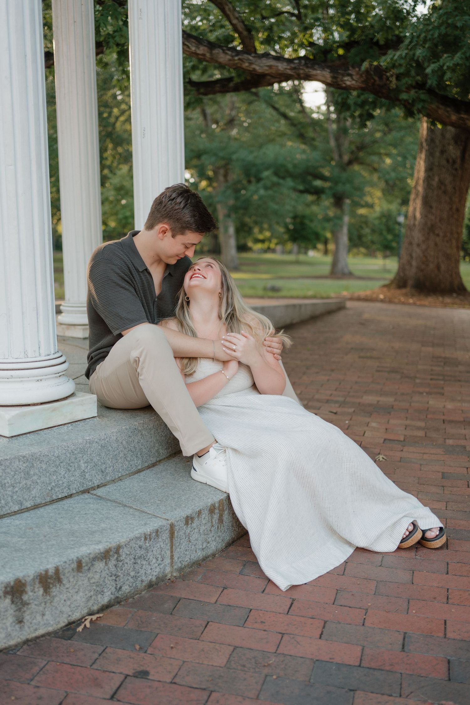 A romantic couple embraces on stone steps beside white columns on a brick pathway surrounded by trees at sunset.