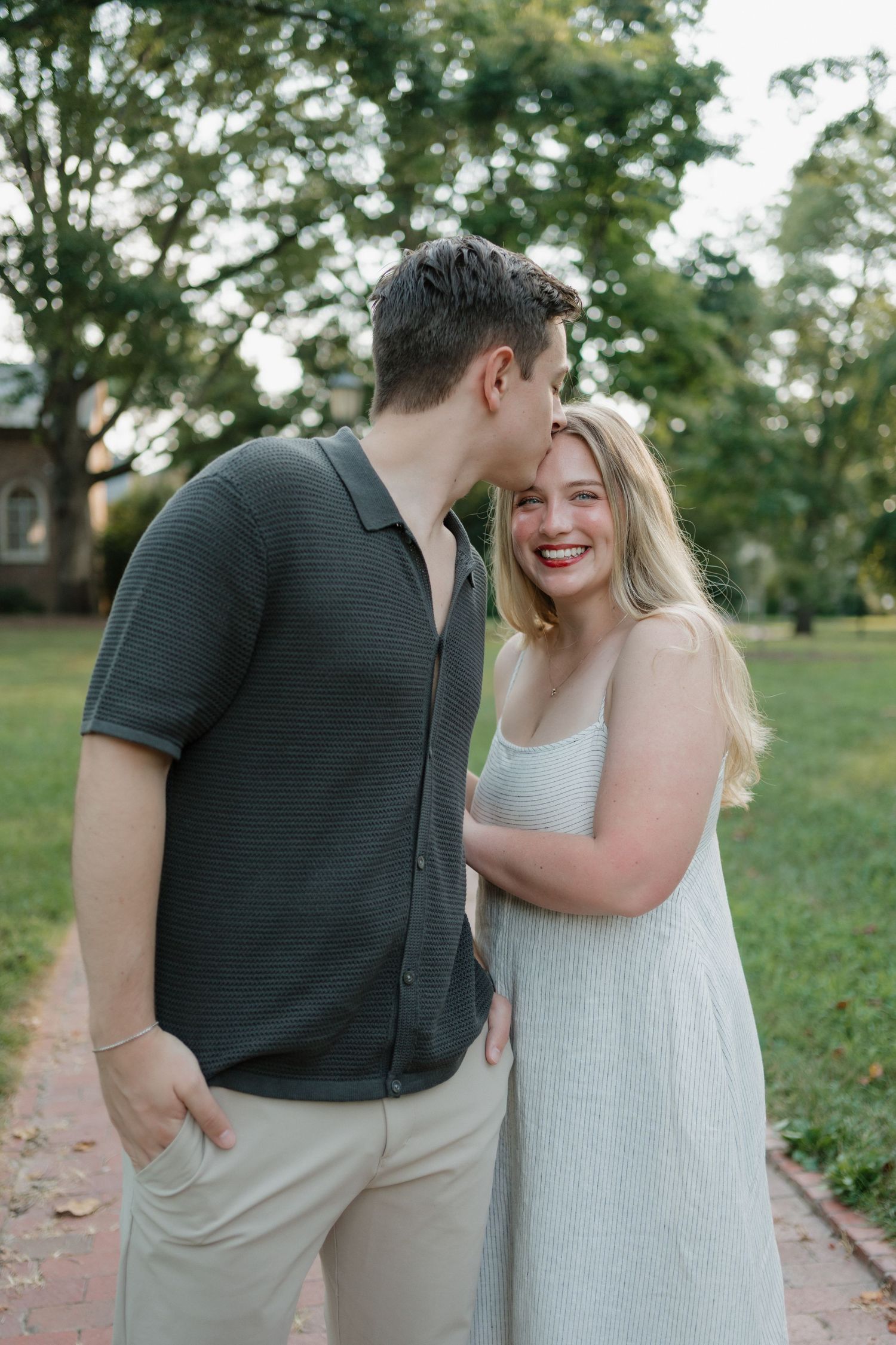 A couple shares an intimate moment together on a brick path surrounded by green grass and trees on a sunny day.