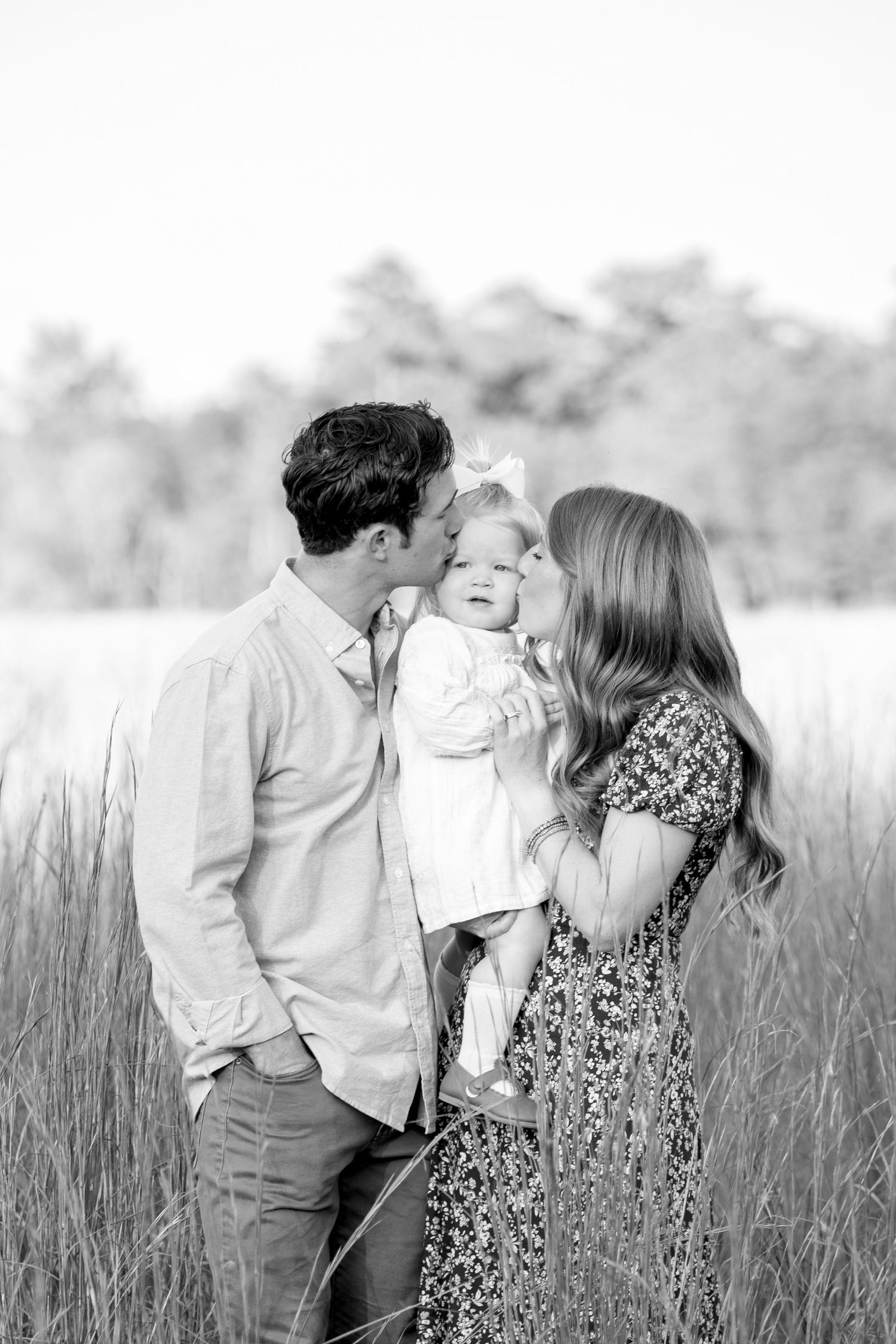 A family shares a tender moment together in a sun-lit field of tall grass, captured in black and white.