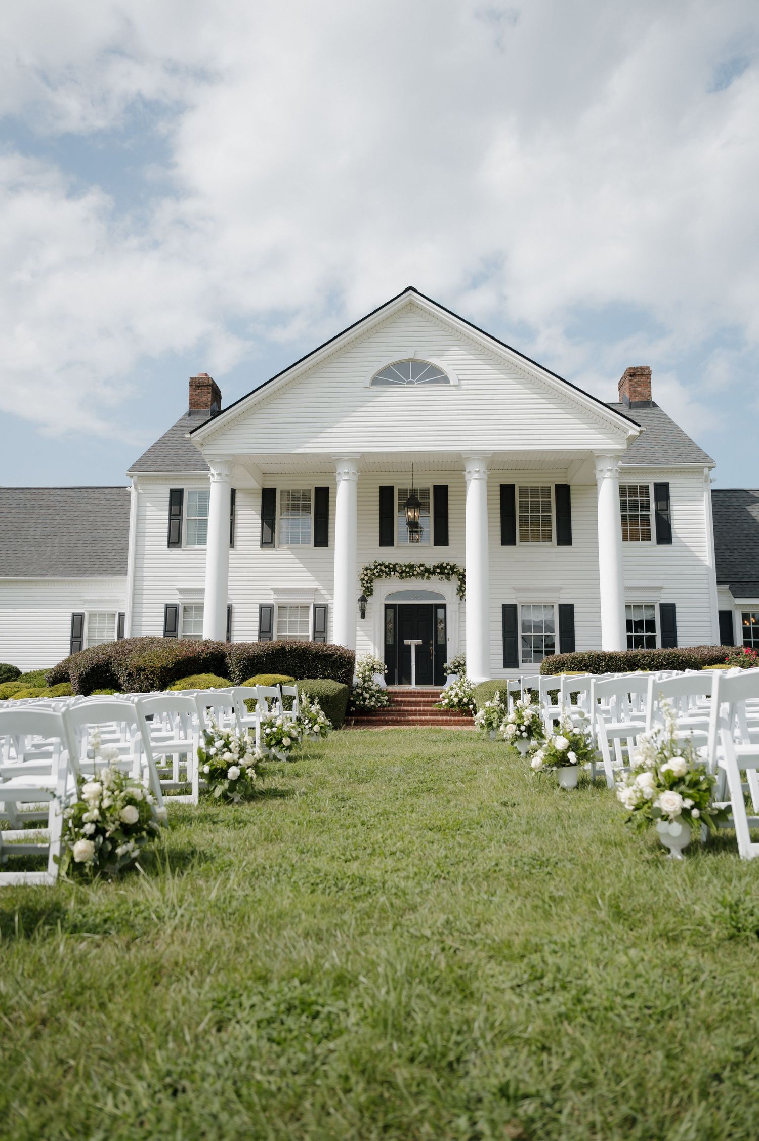 A grand white Southern plantation-style mansion with columns and black shutters set up for an outdoor wedding ceremony.