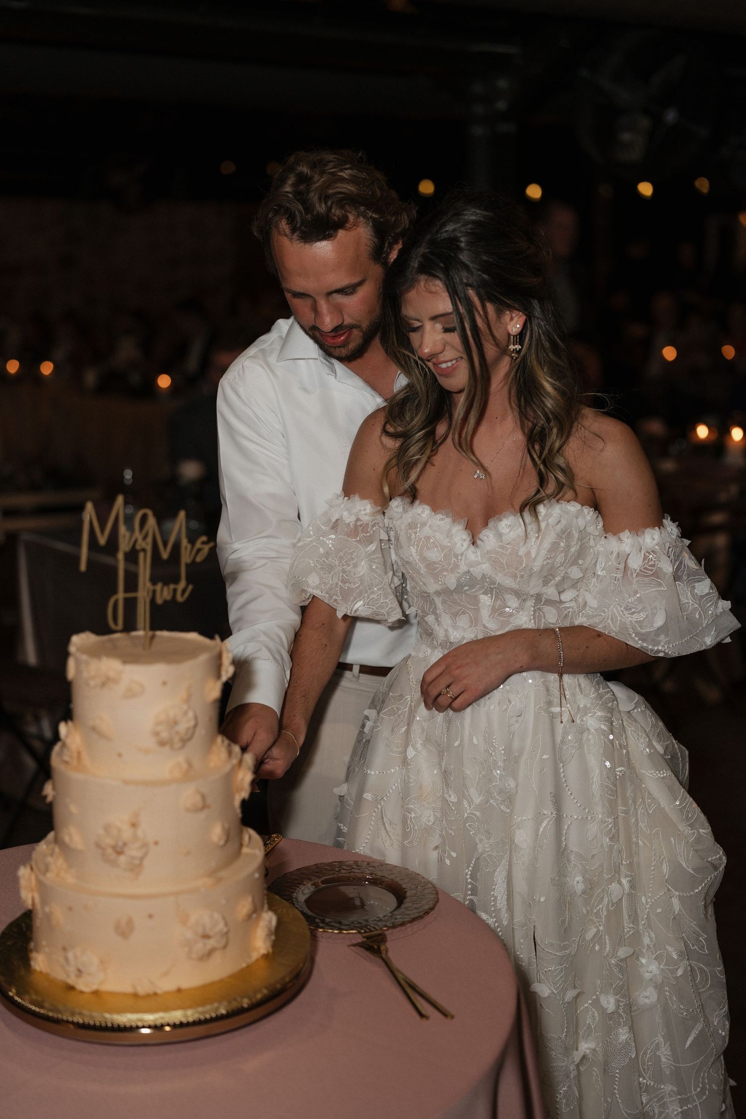 A couple cuts their elegant three-tiered wedding cake decorated with delicate floral details at an evening reception.