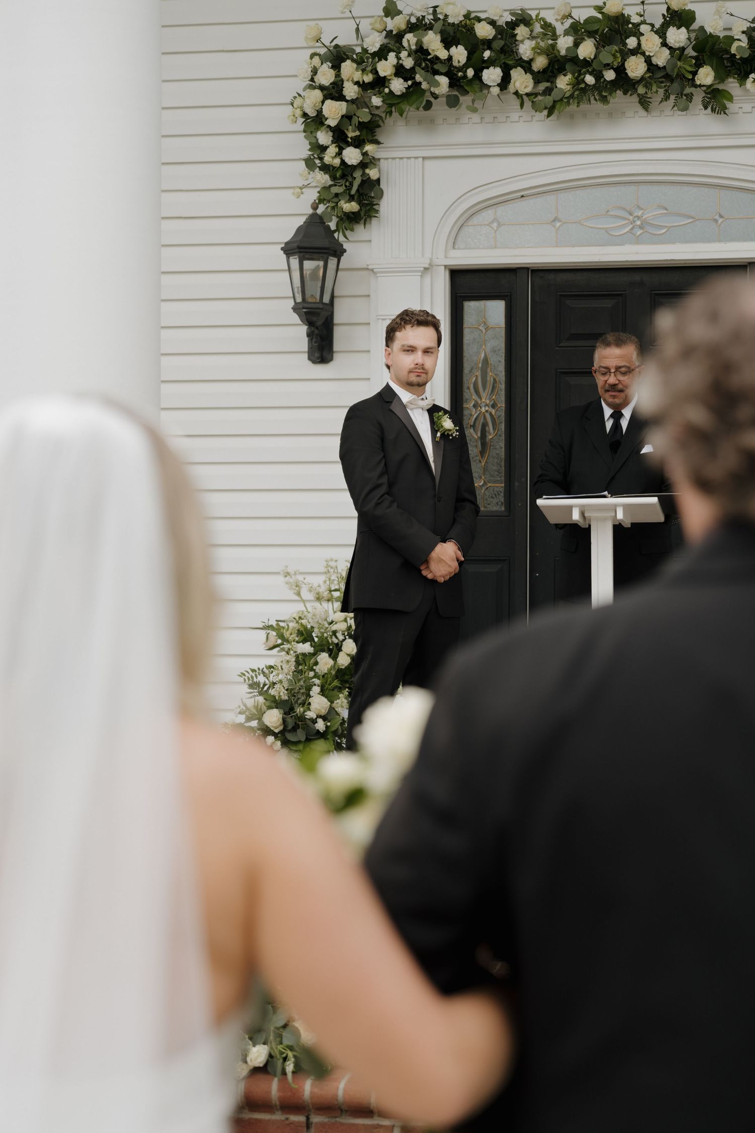 Wedding ceremony with elegant floral arch and black lantern at white colonial entrance.