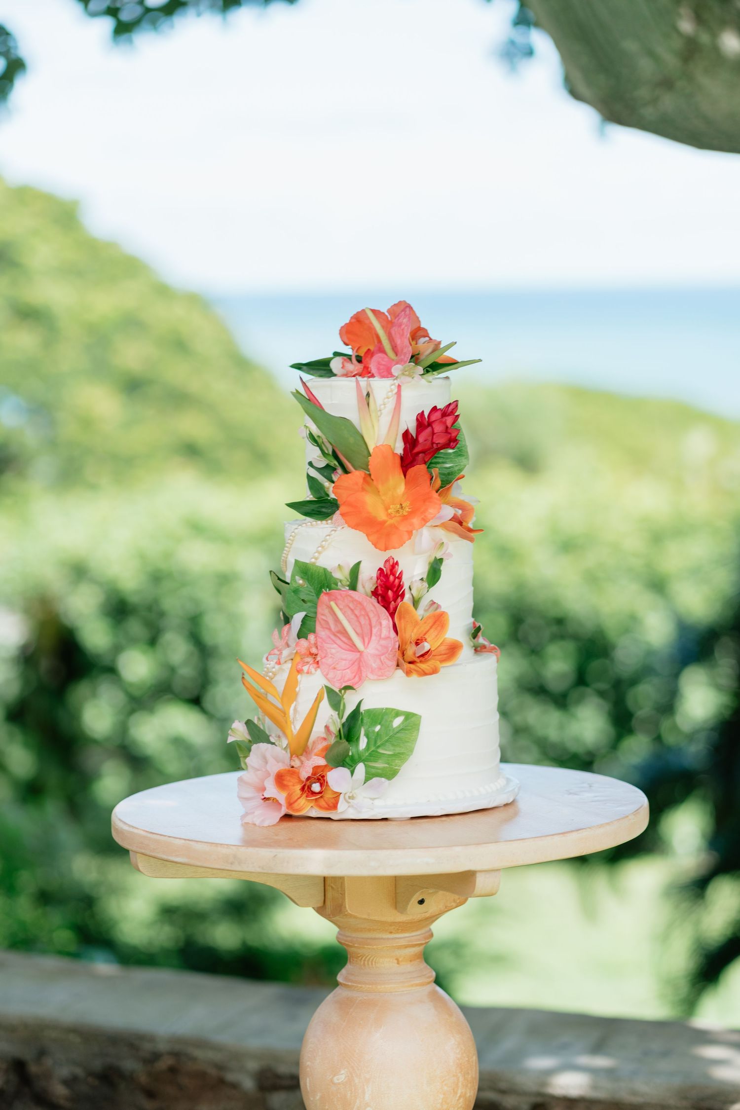 Three-tier white wedding cake adorned with vibrant tropical flowers on a wooden stand with ocean view backdrop.