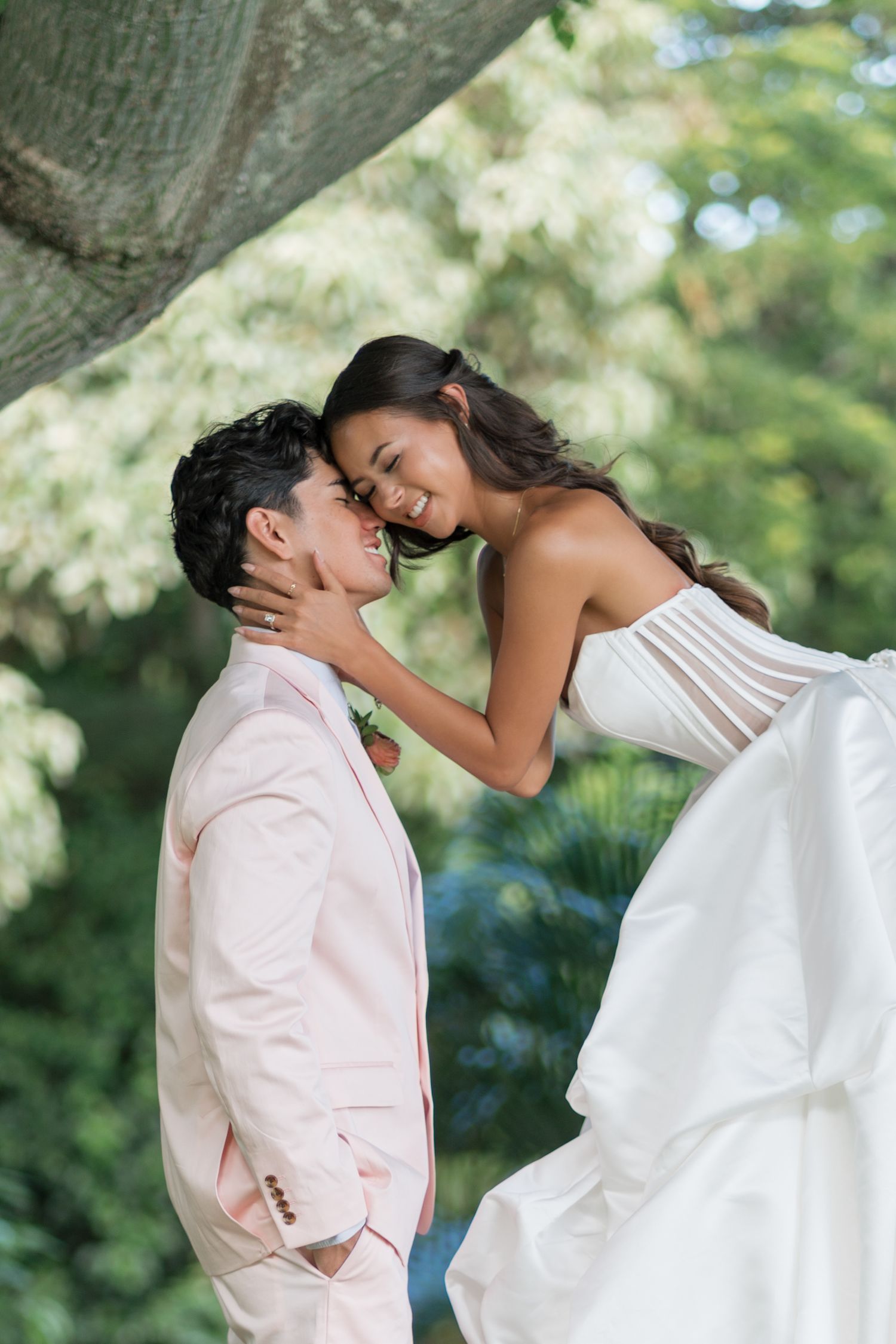 Couple in wedding attire sharing a romantic moment under a tree, bride in white gown, groom in pink suit.