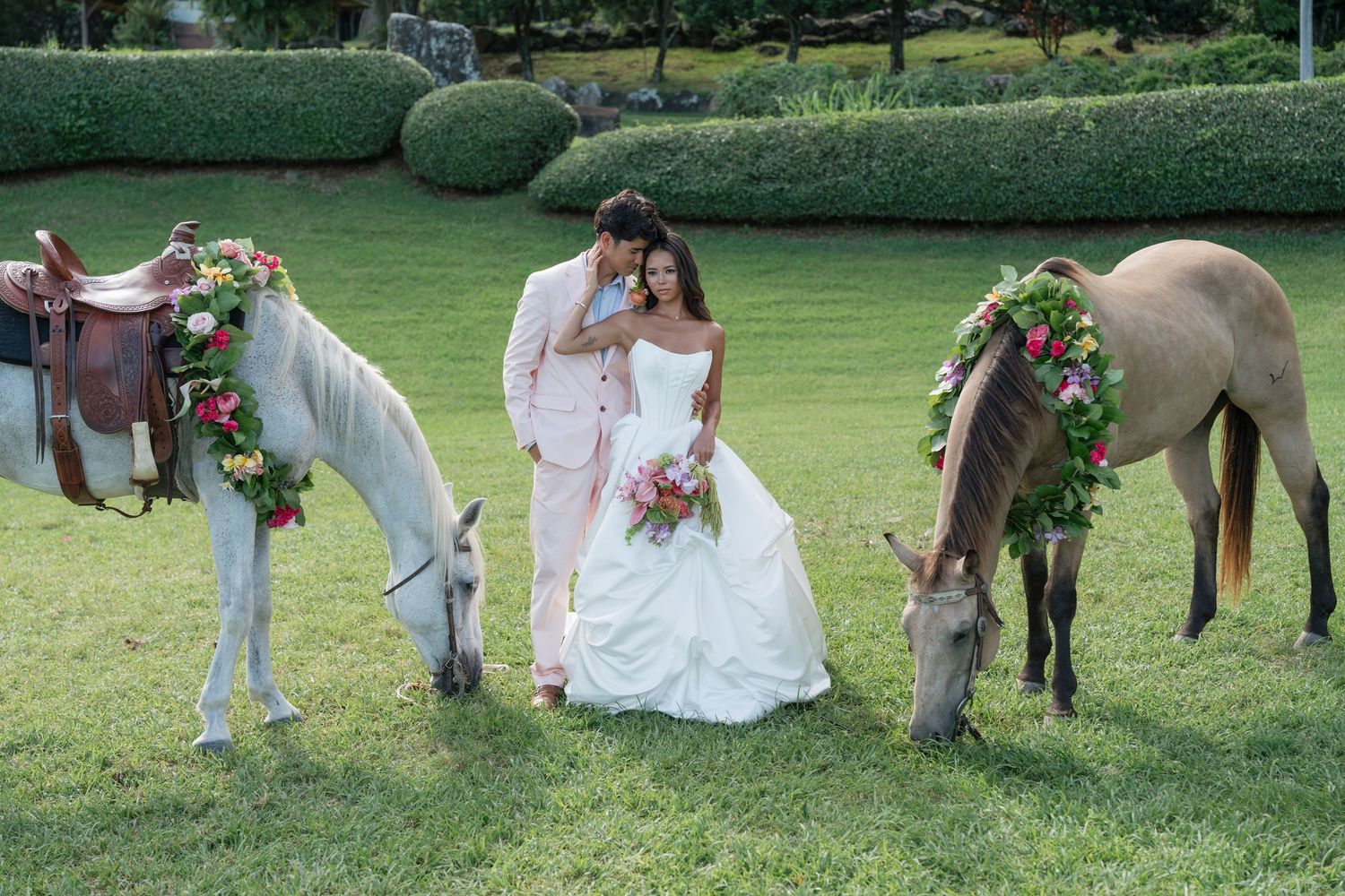 Bride and groom pose between two flower-adorned horses on lush green lawn at outdoor wedding ceremony.