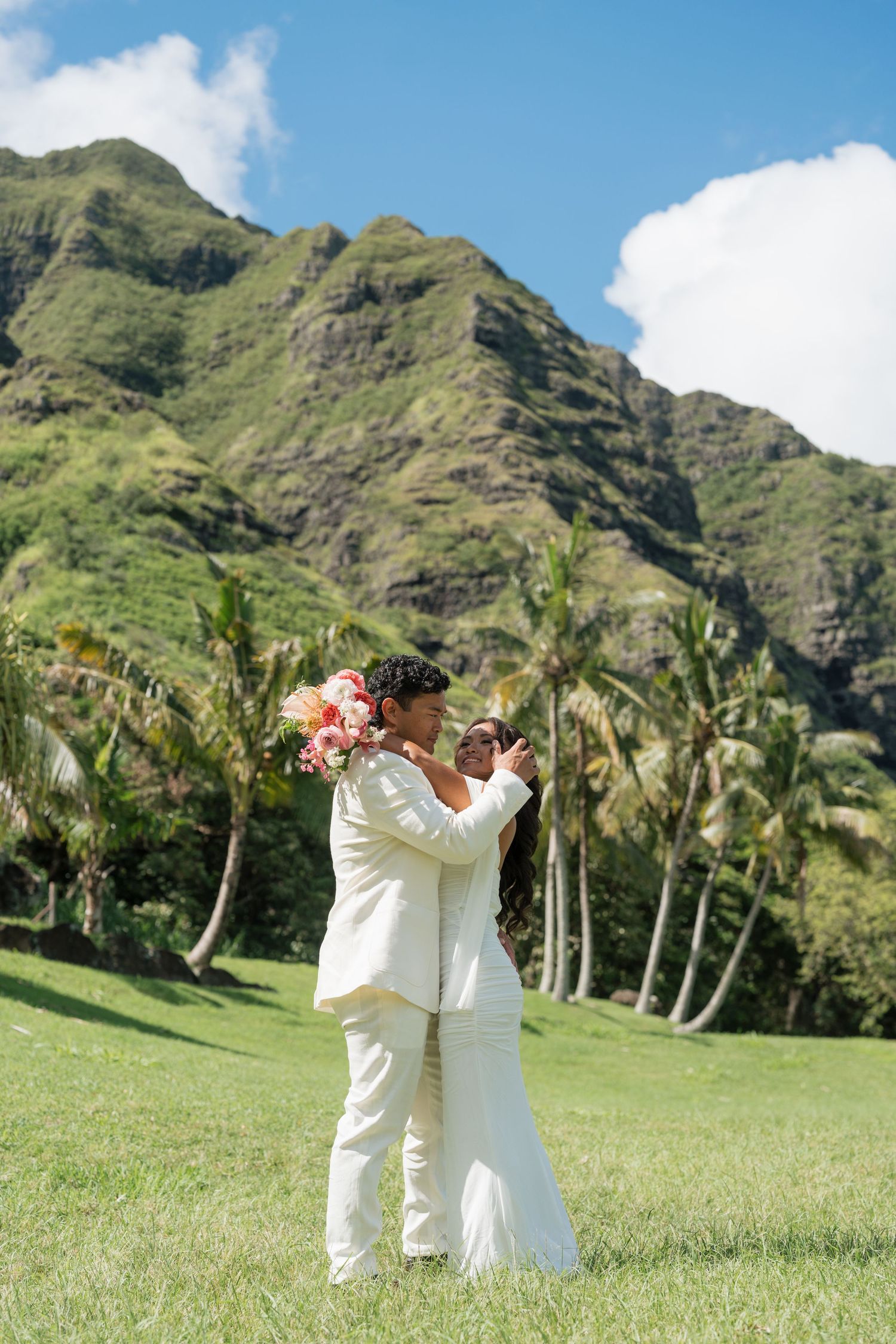 Couple in white wedding attire embracing on green lawn with lush tropical mountains and palm trees in Hawaii.