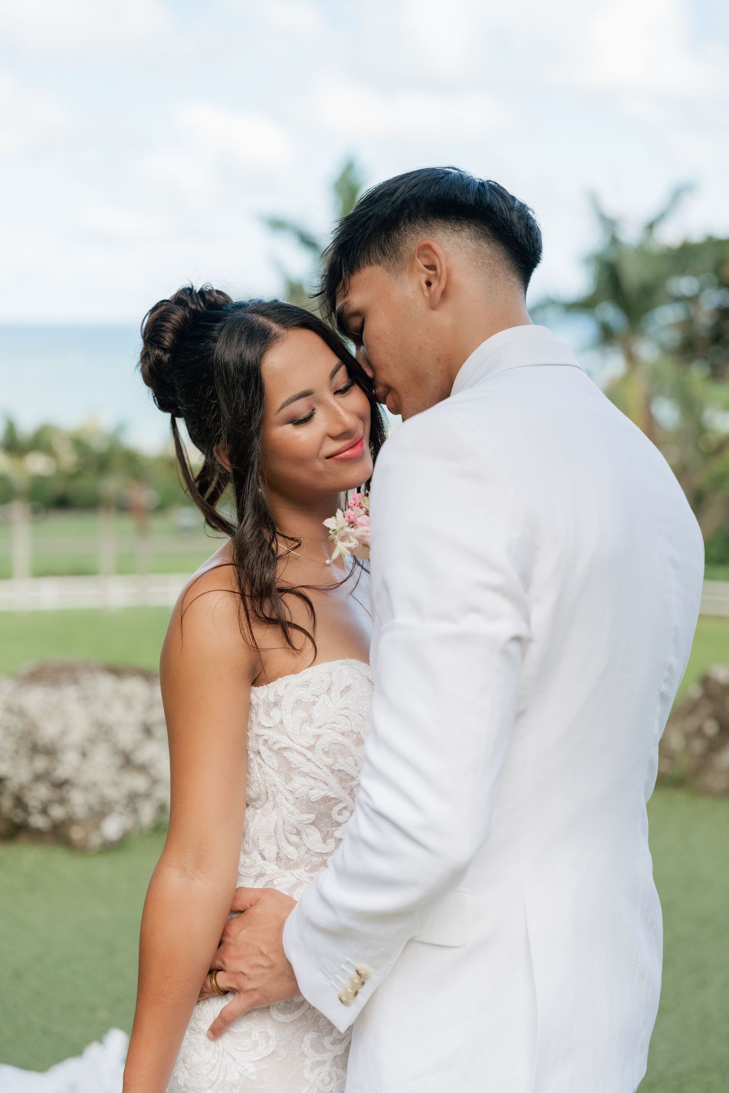 A bride and groom share a romantic embrace outdoors at a tropical wedding venue with palm trees and ocean views.