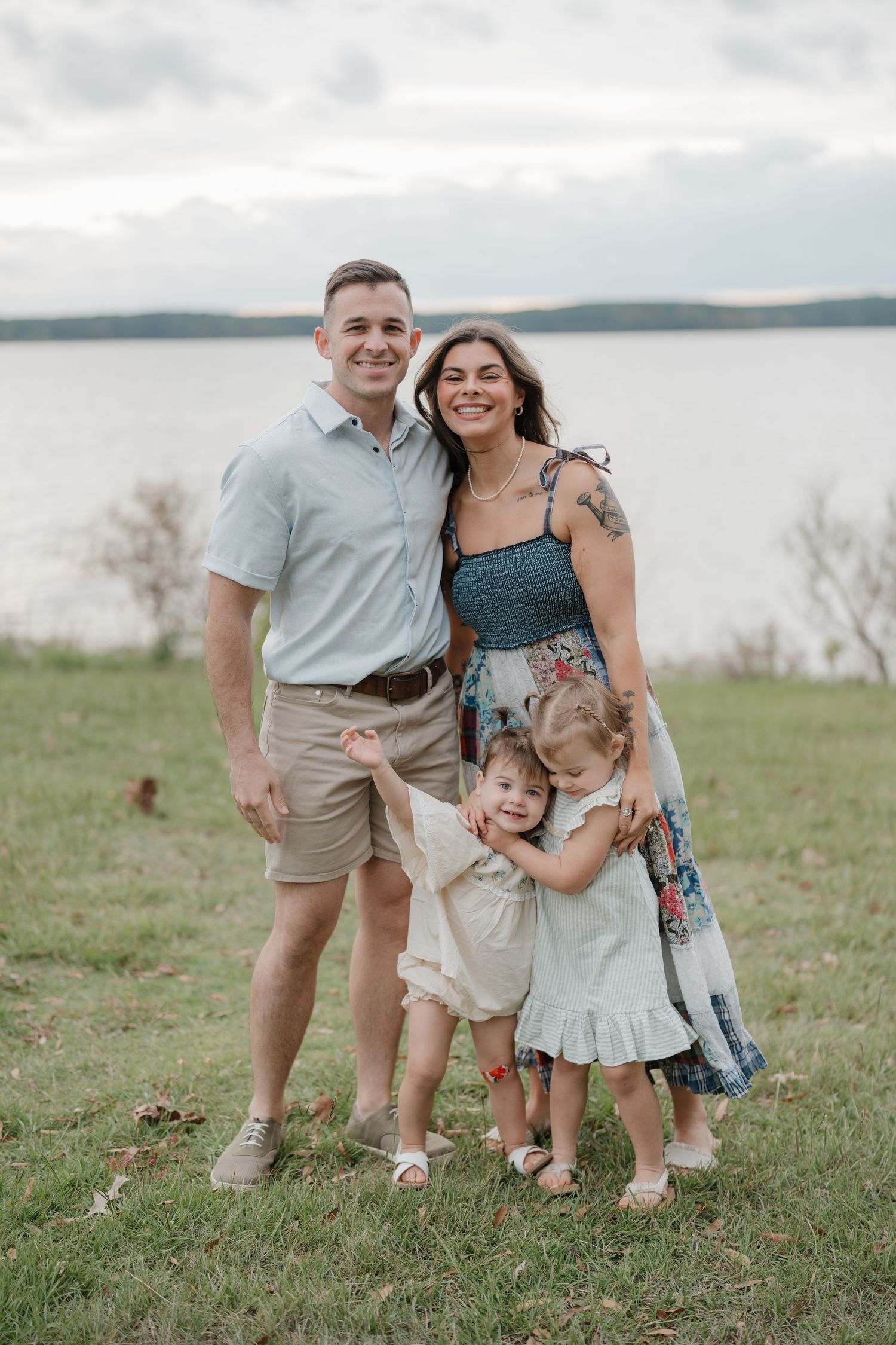 A family stands together on a grassy hill overlooking a serene lake on an overcast day.