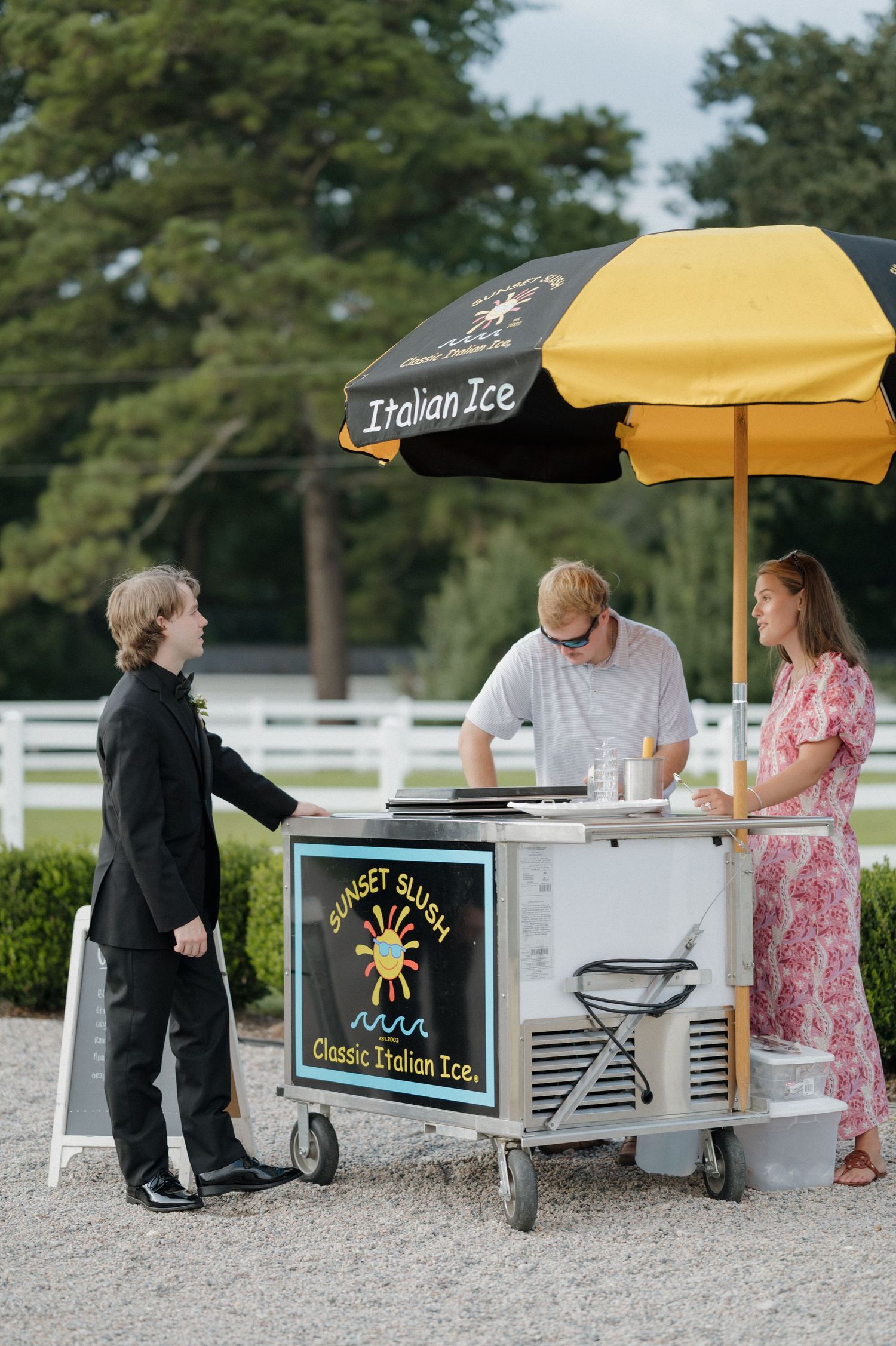 Italian ice vendor serves customers from a mobile cart with black umbrella on a sunny day at an outdoor venue.