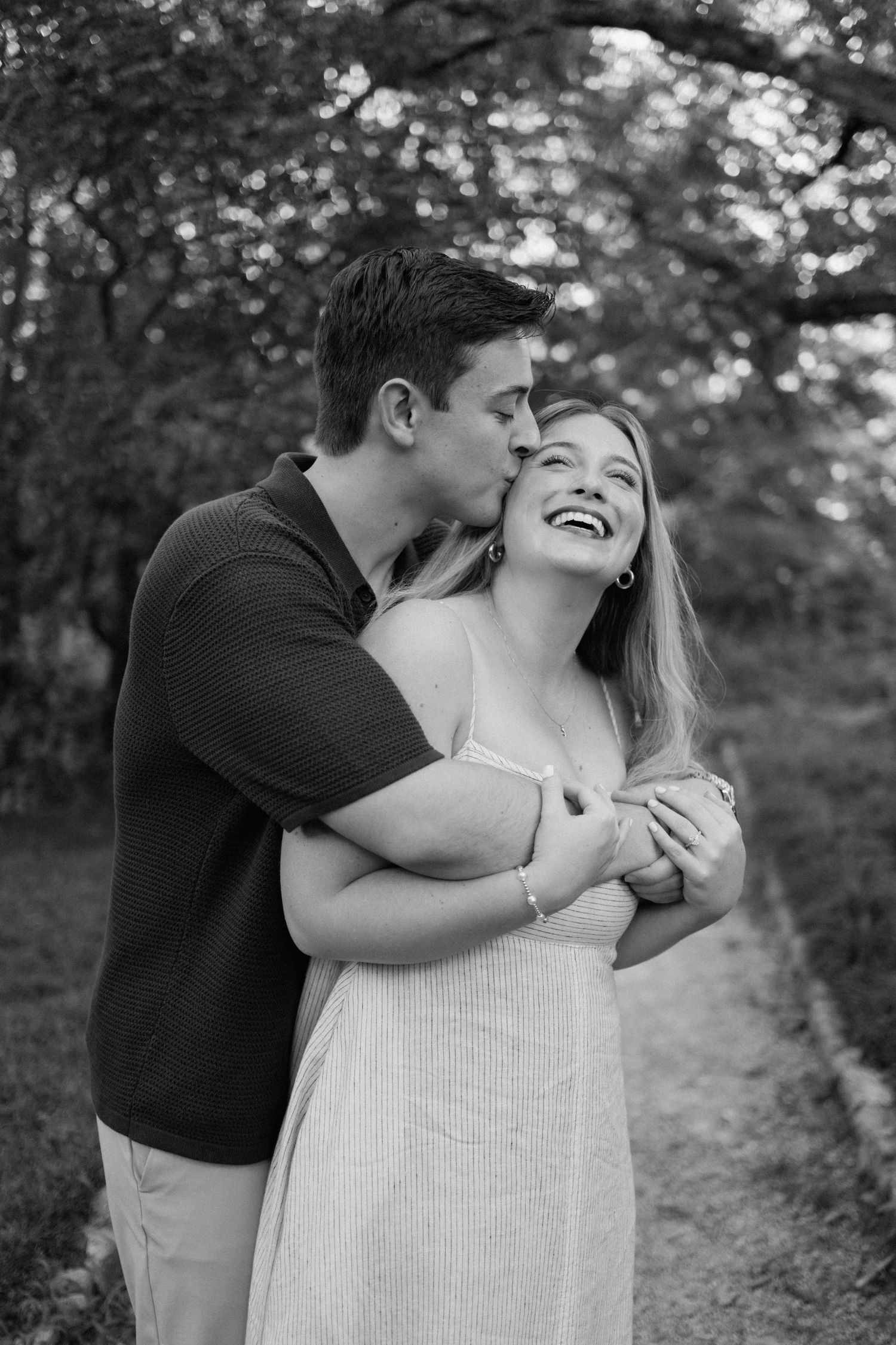 A couple shares a tender moment while laughing together on a tree-lined path in black and white photography.