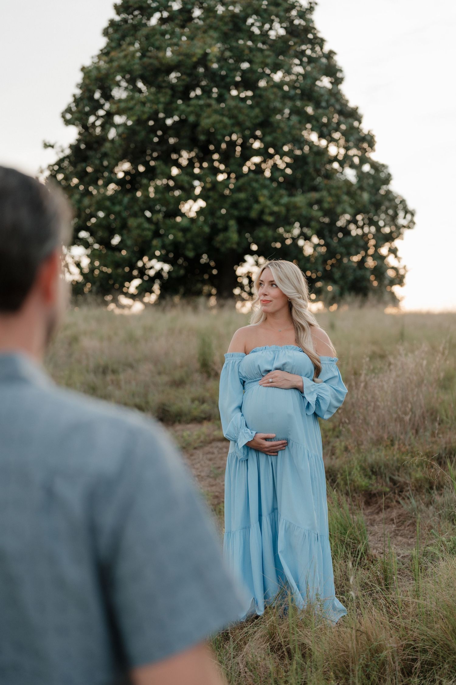 A maternity photoshoot in a grassy field at sunset features an expectant mother wearing a flowing light blue off-shoulder dress.
