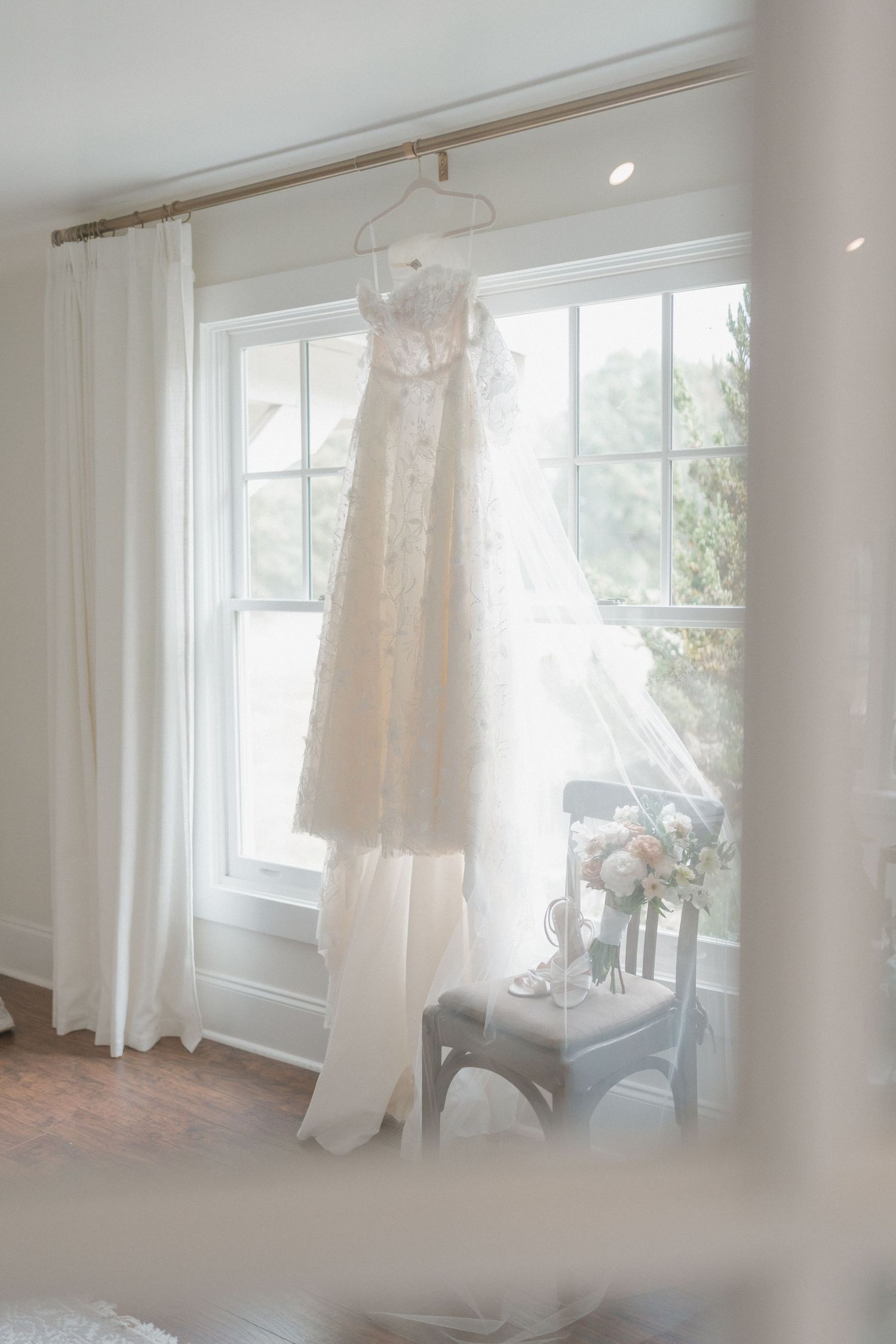 Sunlit bridal preparation room with white curtains and wedding dress hanging by window.