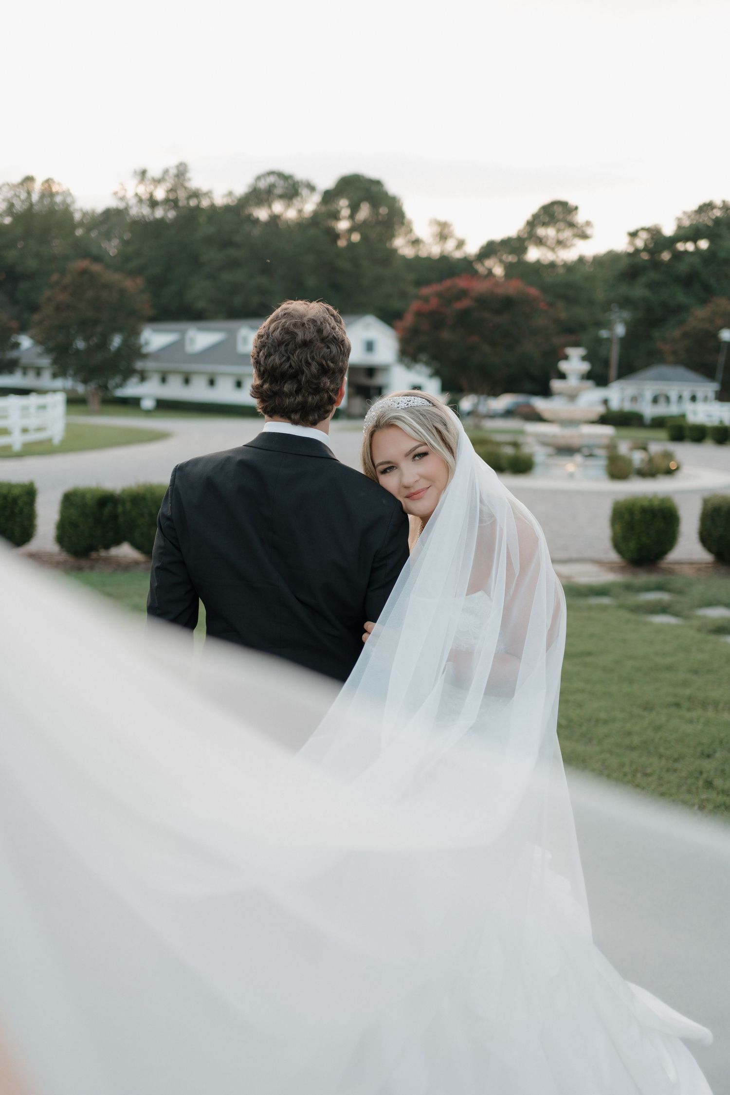 A couple in wedding attire stands on a path with white buildings and trees in the background.
