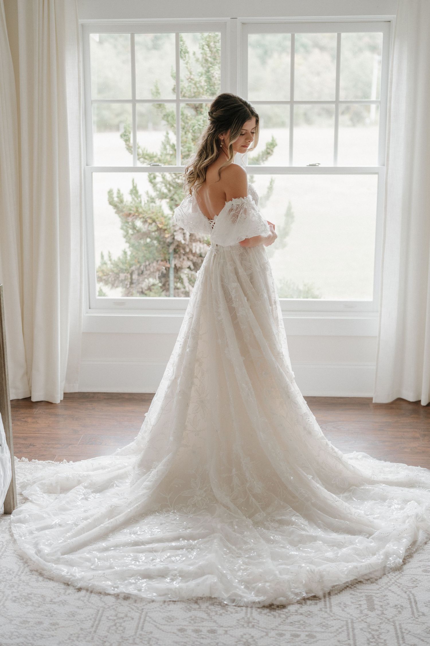 Elegant off-shoulder lace wedding gown with long train photographed in front of bright window with natural light.