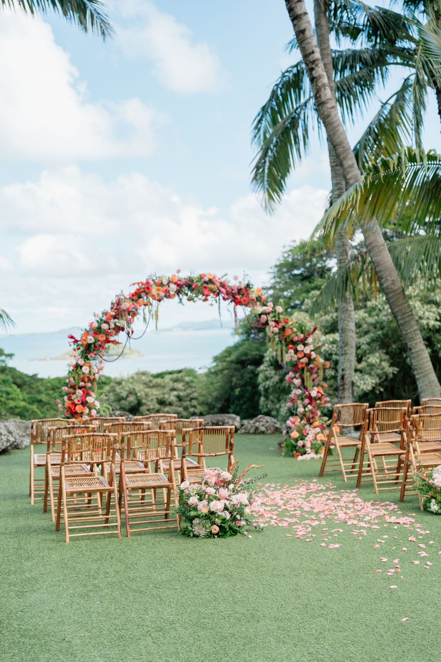 Outdoor wedding ceremony setup with floral arch, wooden chairs, and rose petal aisle on green lawn with palm trees and ocean view.