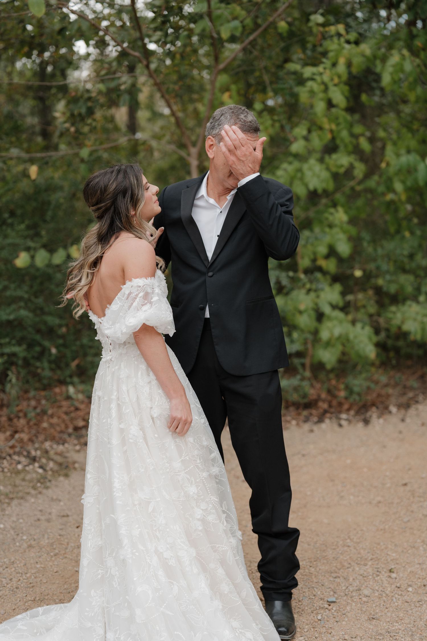 A couple in wedding attire dance together outdoors on a gravel path surrounded by green trees.