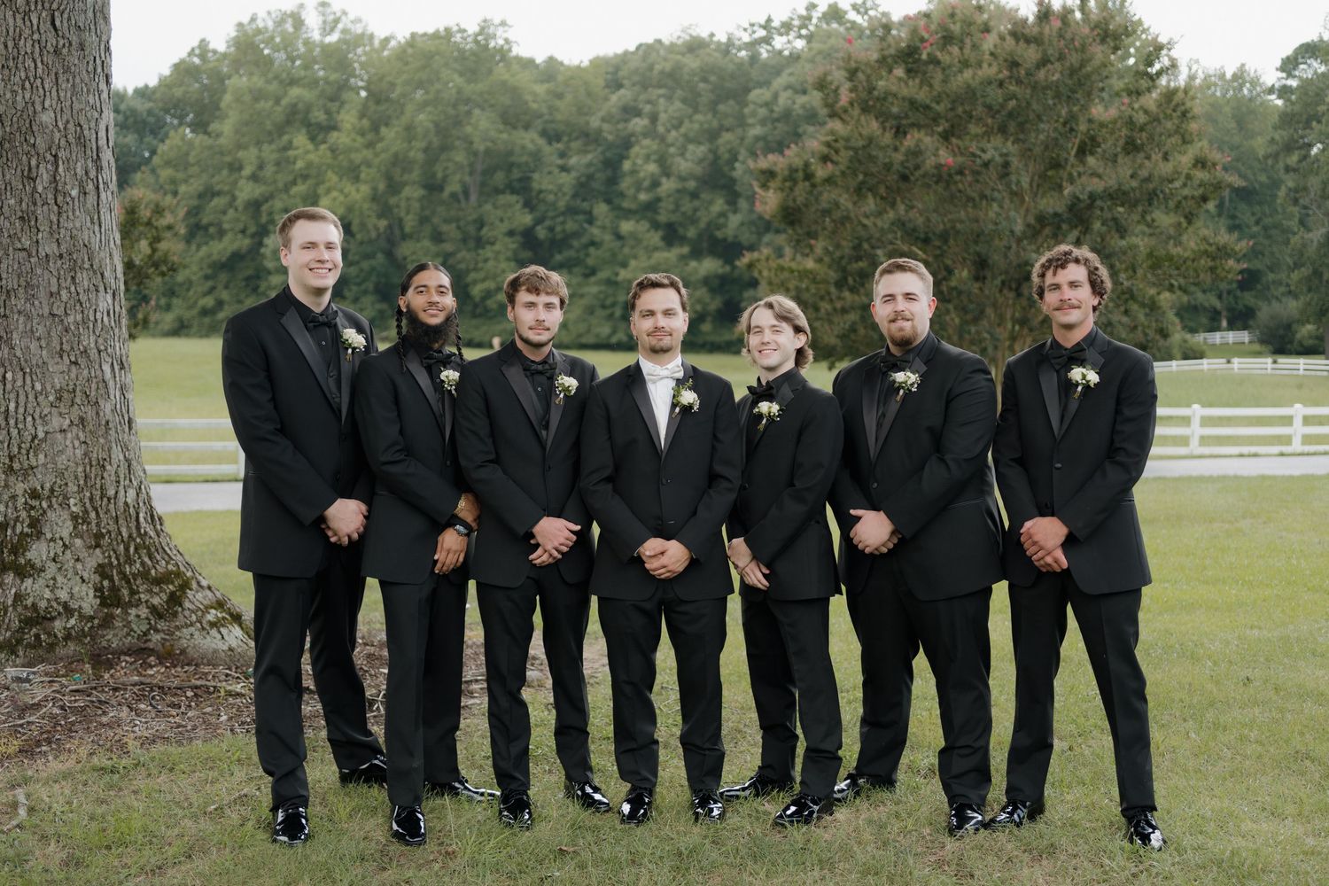 A wedding party in black tuxedos standing in a row on a lawn with white fences and trees in the background.