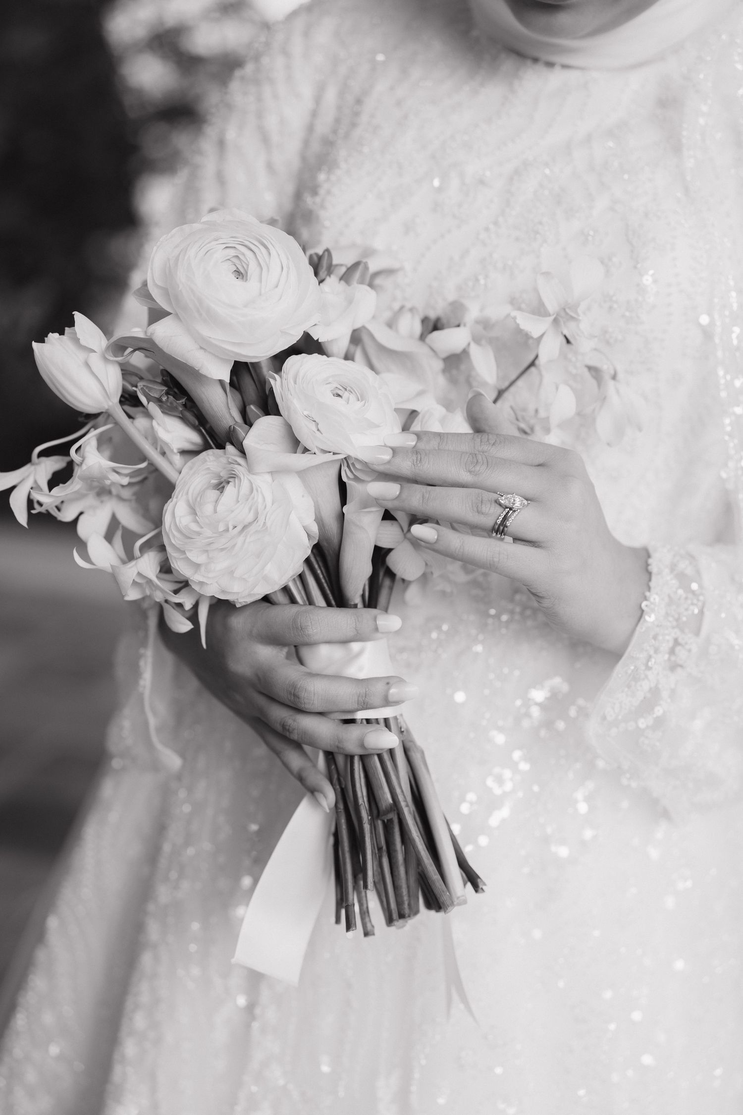Black and white photo of hands holding elegant bridal bouquet with ranunculus flowers and wearing sparkly wedding dress.