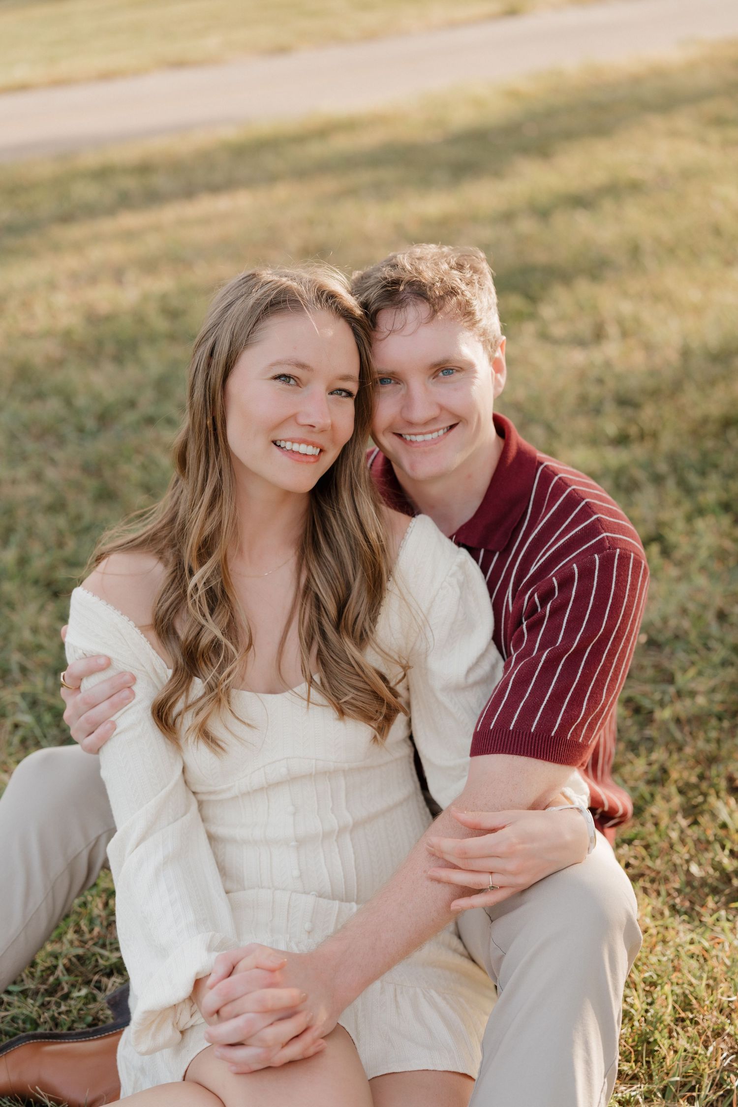 A couple sits together on grass during golden hour, wearing white and burgundy casual attire.