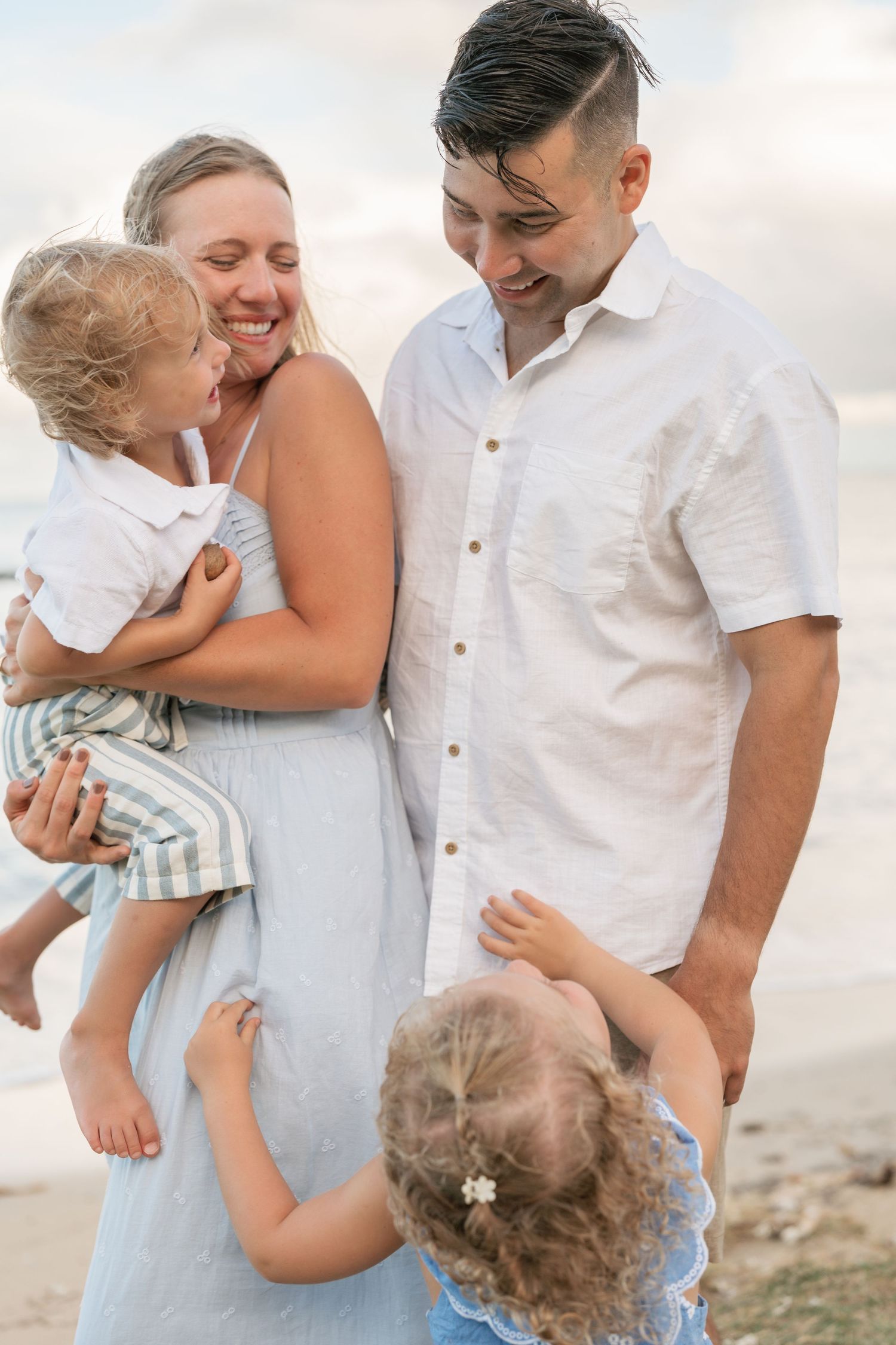 Happy family of four enjoying a beach day, mother holding toddler while daughter hugs parents legs, dressed in light colors.