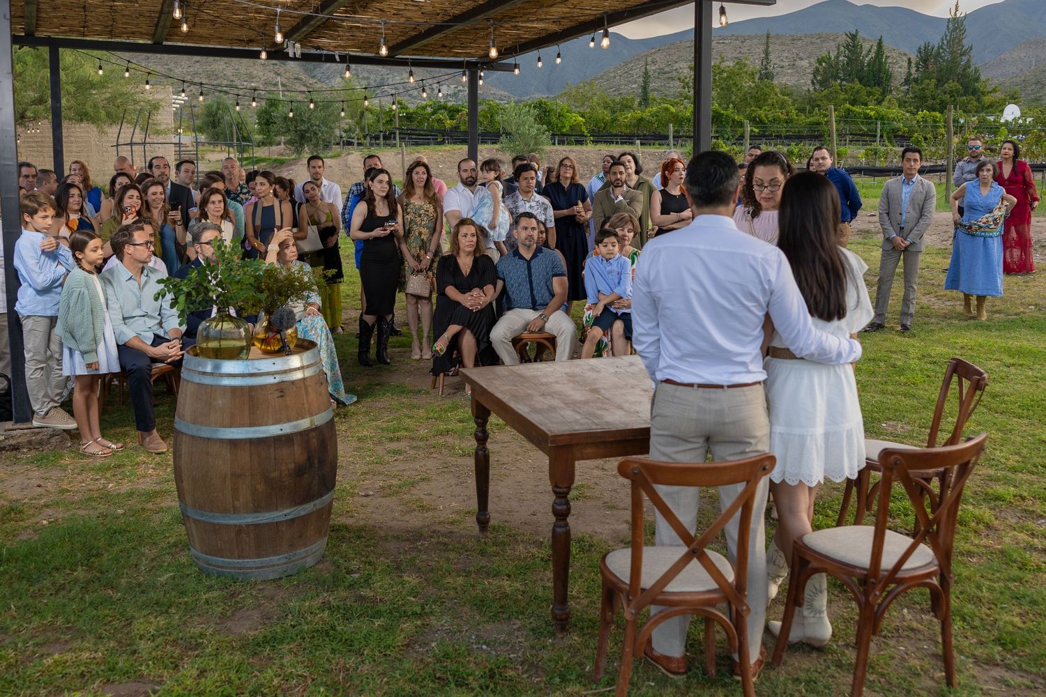 Outdoor celebration setup with wine barrels, wooden furniture and guests gathered under a covered area. Boda en Parras Coahuila fotógrafo