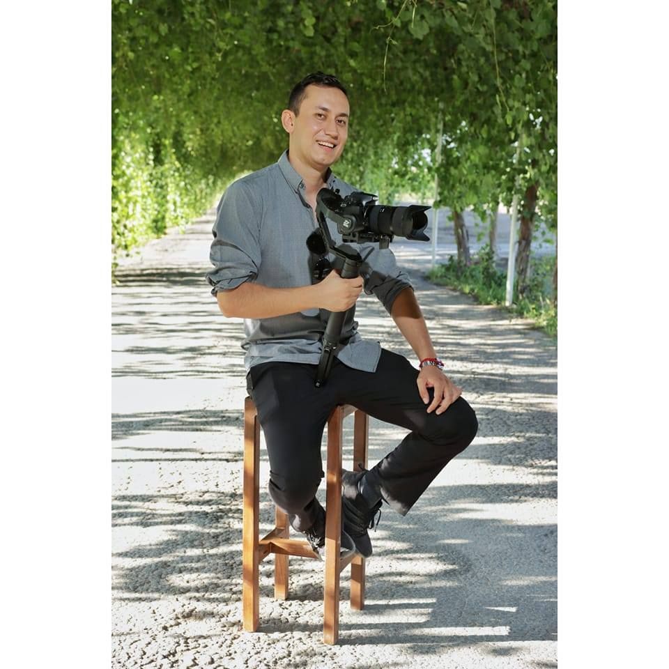 Professional photographer poses with camera gear on wooden stool in outdoor garden setting.