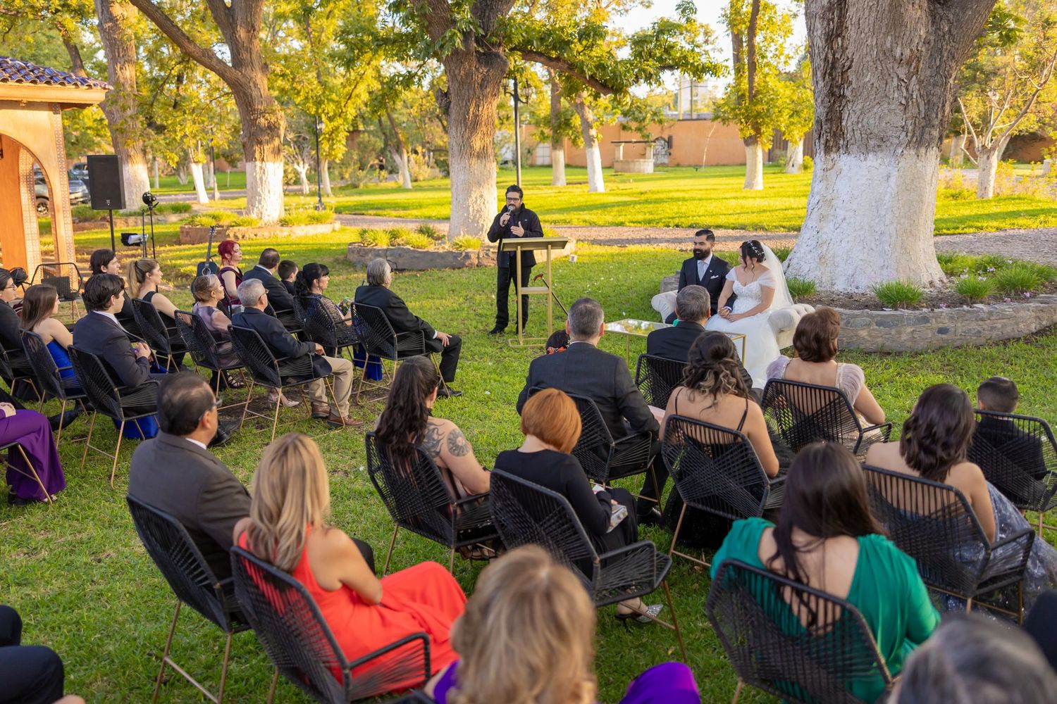 An outdoor wedding ceremony with guests seated on chairs beneath large trees. Boda en Parras Coahuila, Fotógrafo de bodas