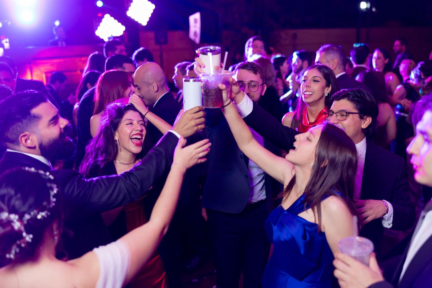 Celebration on dance floor with guests raising drinks under purple party lighting. Boda en Parras Coahuila, Fotógrafo de bodas