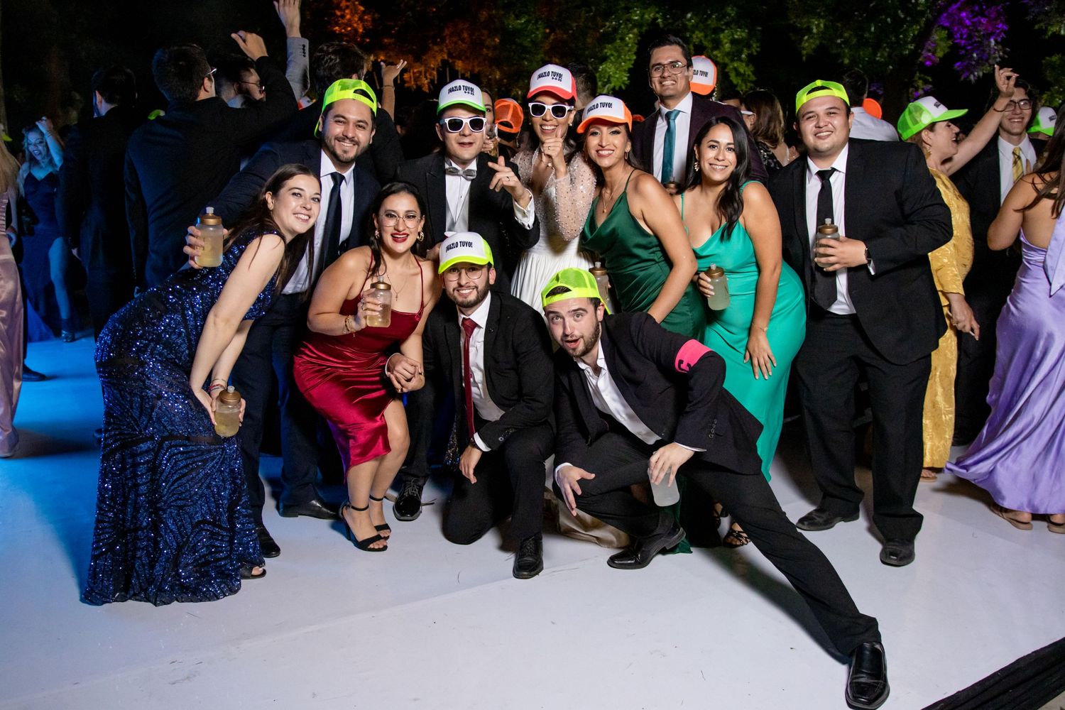 Wedding guests in formal attire pose together at an outdoor evening reception wearing festive hats. Boda en Parras Coahuila, Fotógrafo