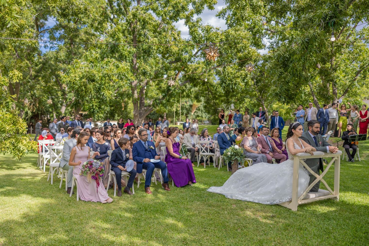 Large wedding gathering with guests seated on white chairs under shaded trees. Boda en Parras Coahuila fotógrafo