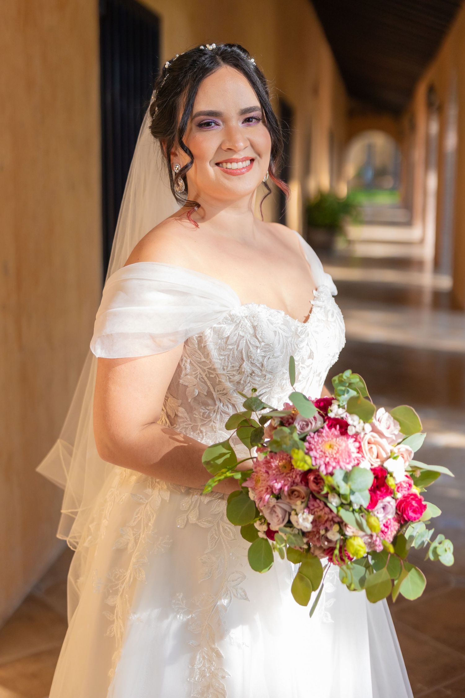 A bride in an off-shoulder wedding gown holds a colorful bouquet in a Spanish-style corridor. Boda en Parras Coahuila, Fotógrafo de bodas