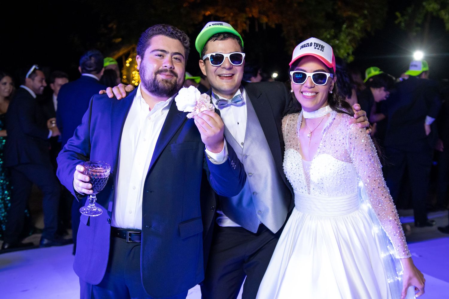 Guests celebrate at a nighttime wedding reception wearing colorful formal attire and baseball caps. Boda en Parras Coahuila, Fotógrafo