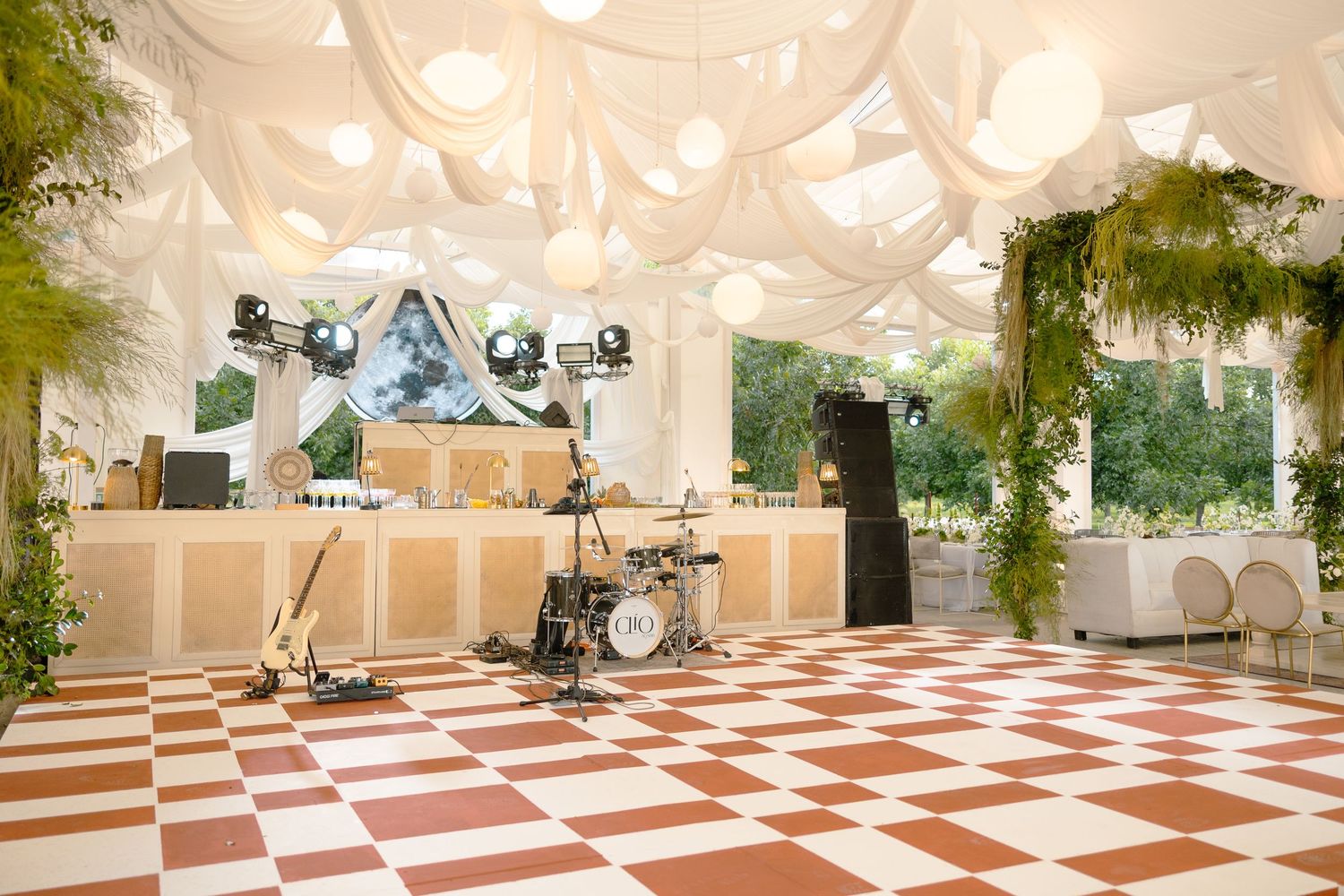 A luxurious event venue with white draping, checkerboard dance floor, and elegant decor featuring hanging greenery.