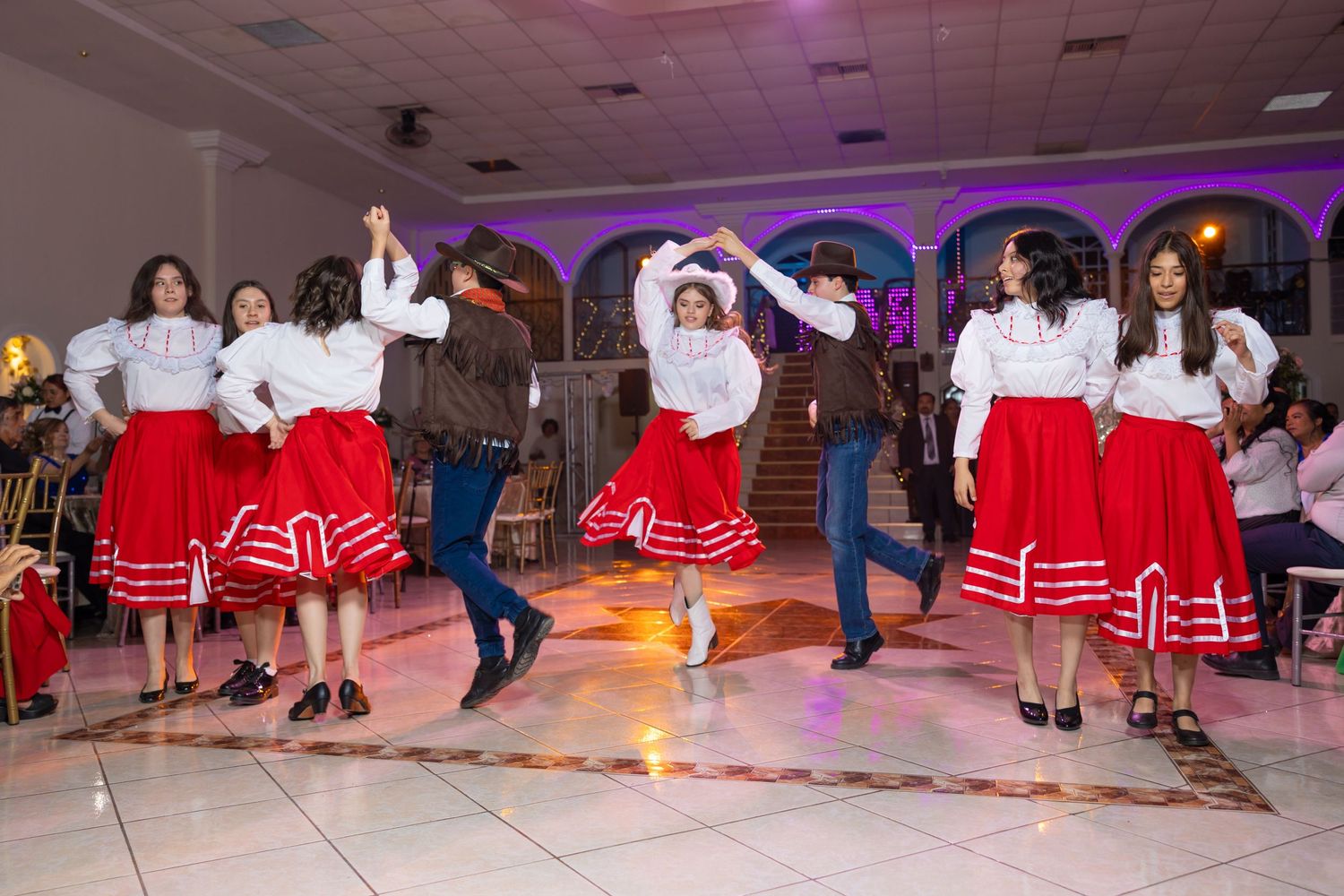 Dancers in white tops and red skirts perform synchronized moves under purple venue lighting. Evento en Parras Coahuila , Fotógrafo Rulo