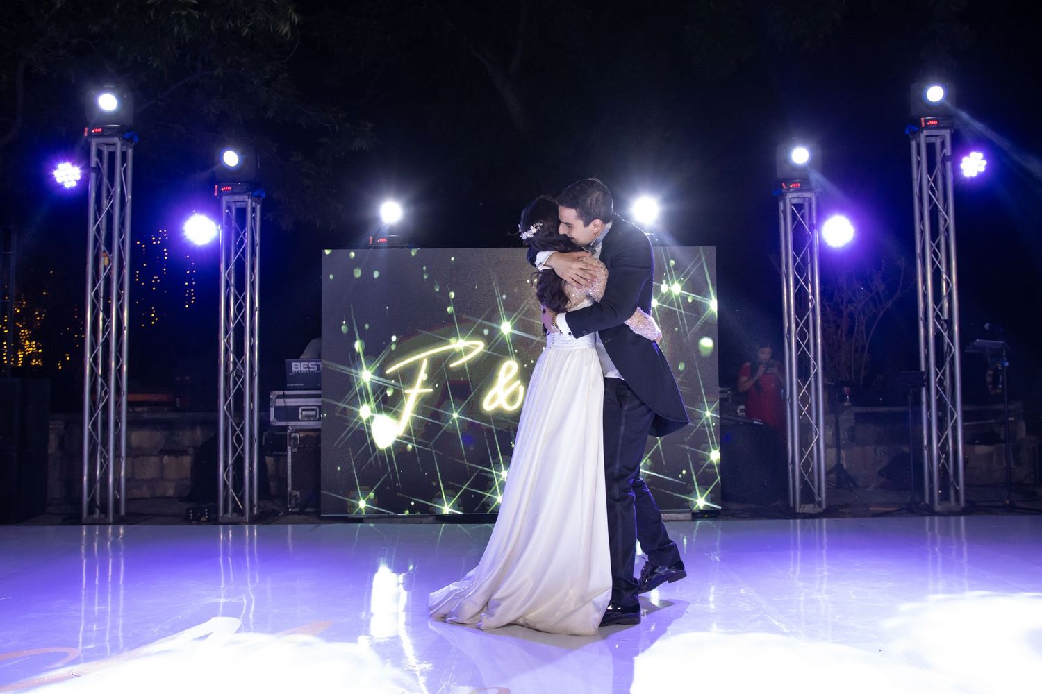 A couple shares their first dance on an illuminated stage with blue lighting and truss structures. Boda en Parras Coahuila, Fotógrafo
