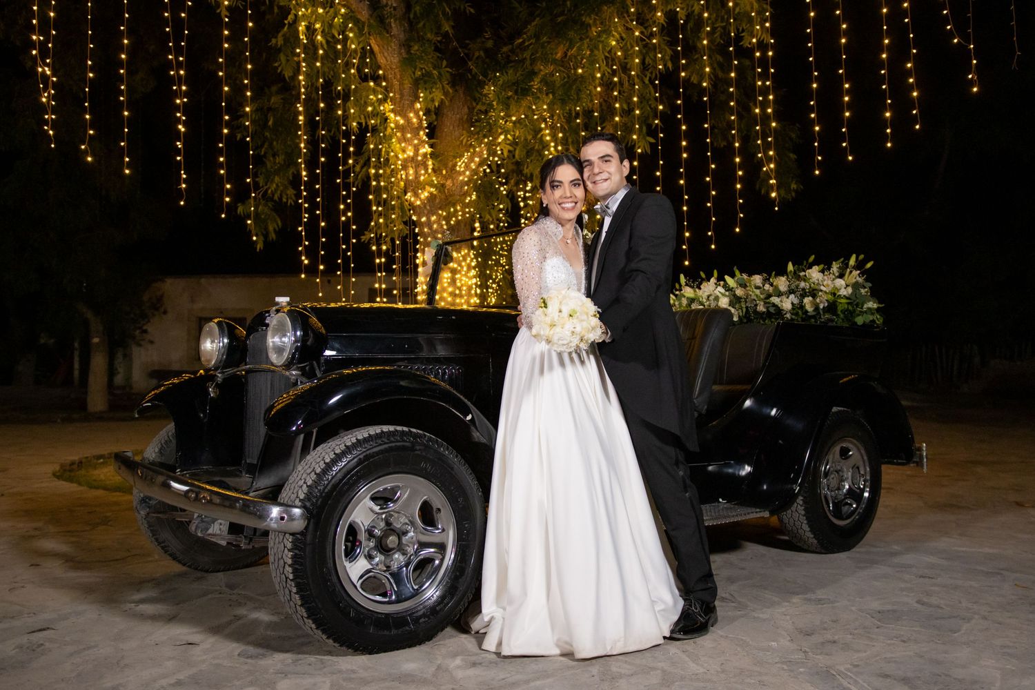 Wedding couple poses with vintage black car under romantic string lights. Boda en Parras Coahuila, Fotógrafo de bodas