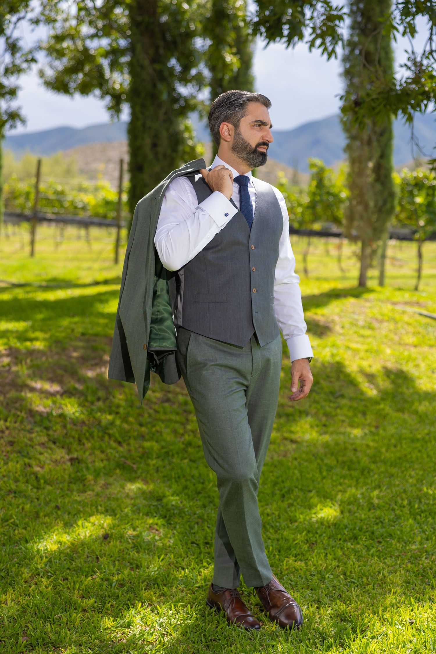 Stylish man in business attire poses with jacket draped over shoulder in scenic vineyard landscape. Boda en Parras Coahuila fotógrafo