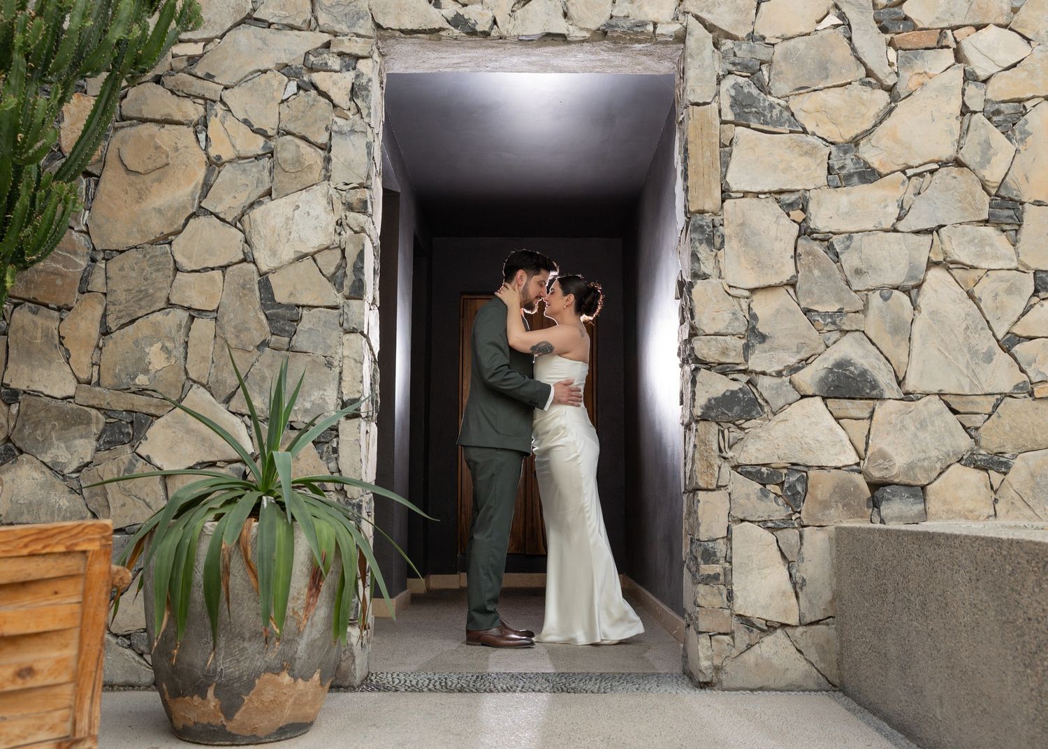 A bride in a white wedding dress walks past a rustic stone wall with potted plants and wooden furniture.