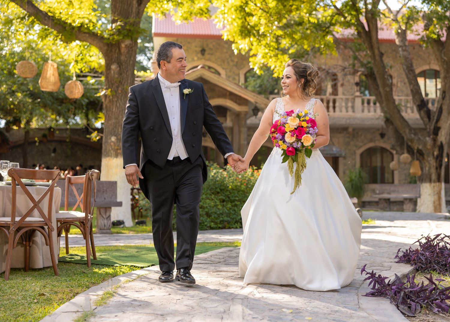 Bride and groom walk hand in hand through a sunlit garden path with golden autumn leaves overhead.