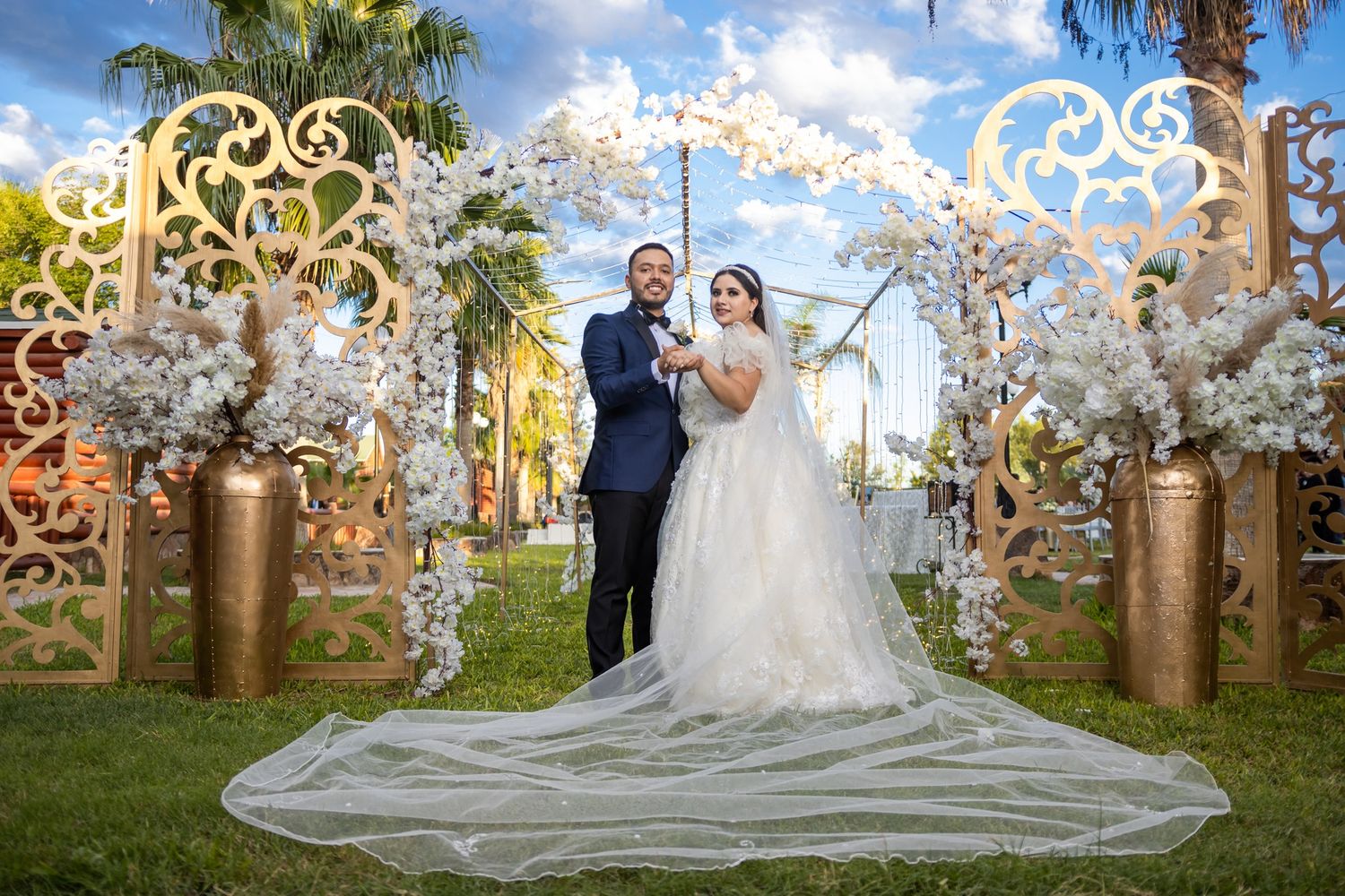 An elegant wedding arch decorated with white flowers and golden accents frames a couple in a garden setting. Boda en Parras Coahuila fotógra