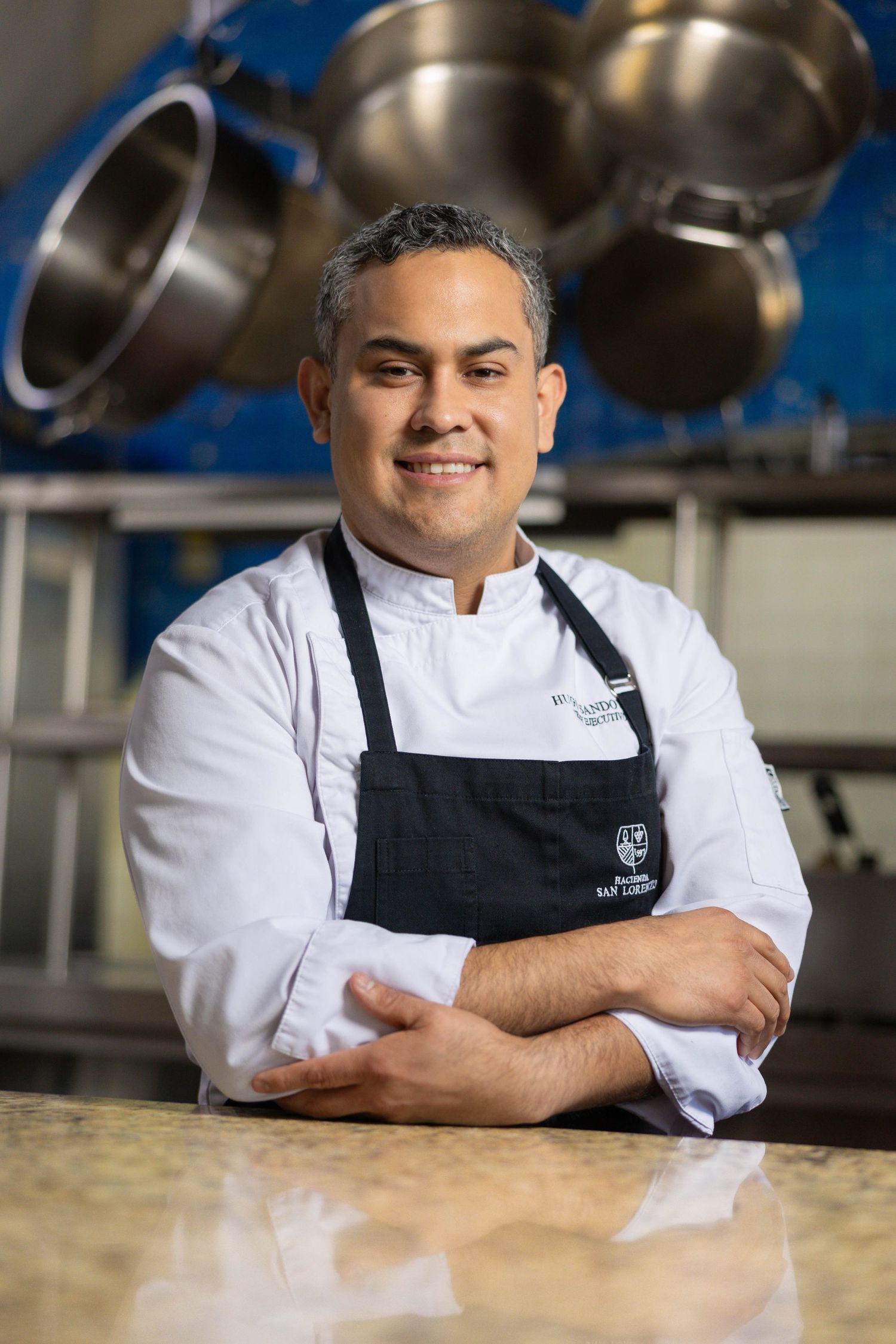A chef in white coat and black apron stands confidently in a professional kitchen with cookware in the background.