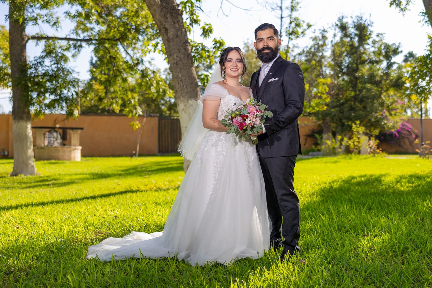 A wedding couple poses together on a sunny lawn with trees and architecture in the background. Boda en Parras Coahuila, Fotógrafo de bodas