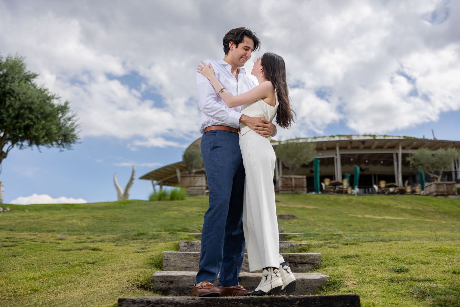 A romantic couple embraces on steps in a grassy field under cloudy skies. Propuesta de matrimonio Parras, Fotografo