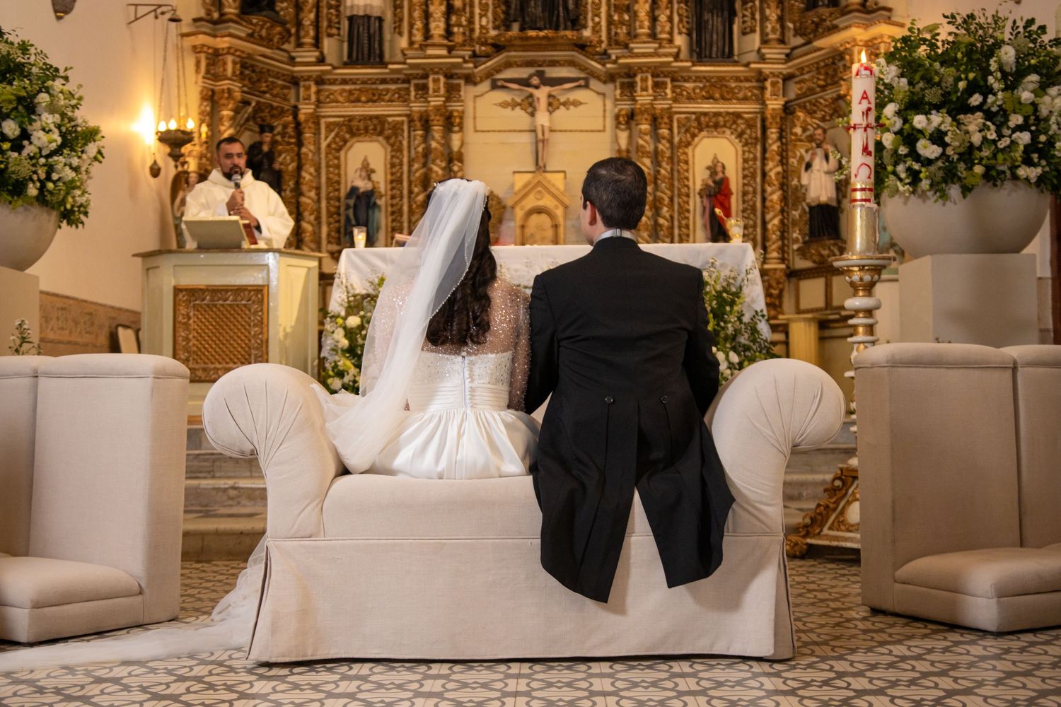 Couple seated on white couch before ornate golden altar during ceremony. Boda en Parras Coahuila, Fotógrafo de bodas