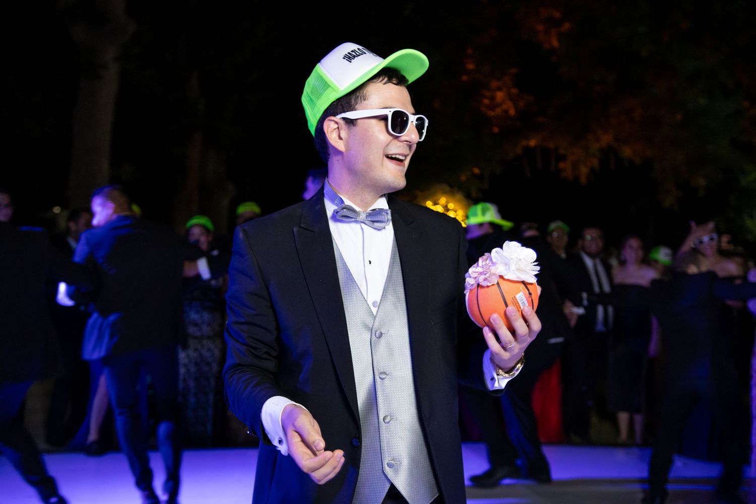 A guest in formal attire and neon baseball cap enjoys refreshments at an outdoor wedding reception. Boda en Parras Coahuila, Fotógrafo