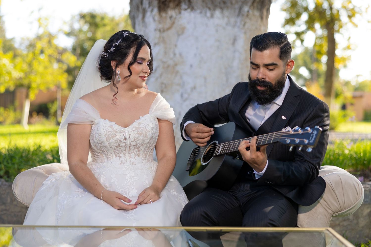 A couple sits on a bench outdoors while one plays guitar in wedding attire. Boda en Parras Coahuila, Fotógrafo de bodas