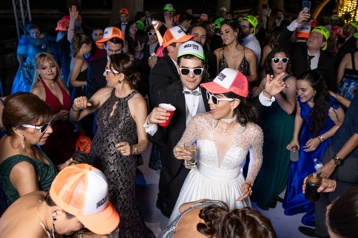 Wedding party guests dance and celebrate on a crowded dance floor wearing colorful formal attire and festive hats. Boda en Parras Coahuila