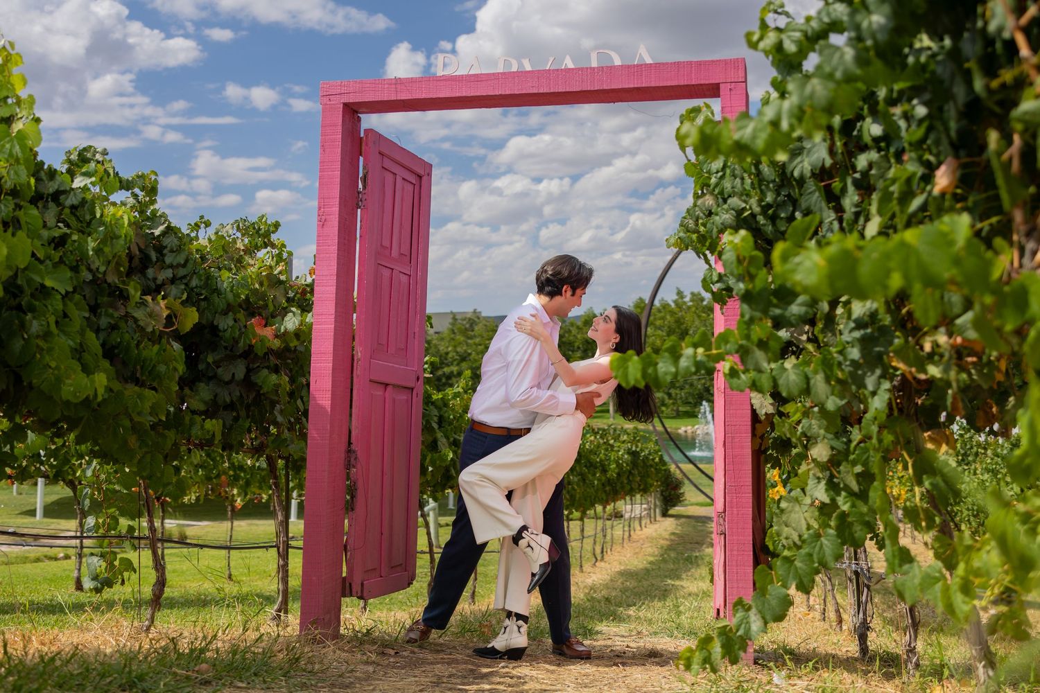 A couple shares an intimate moment beneath a pink doorway in a sunny vineyard. Propuesta de matrimonio Parras, Fotografo