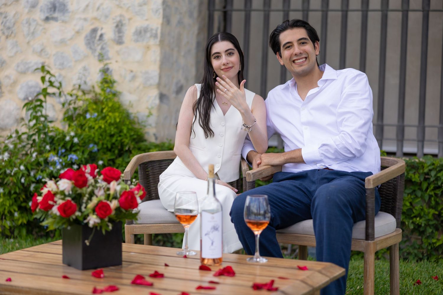 A couple seated outdoors shares a moment among red flowers and garden decor. Propuesta de matrimonio Parras, Fotografo