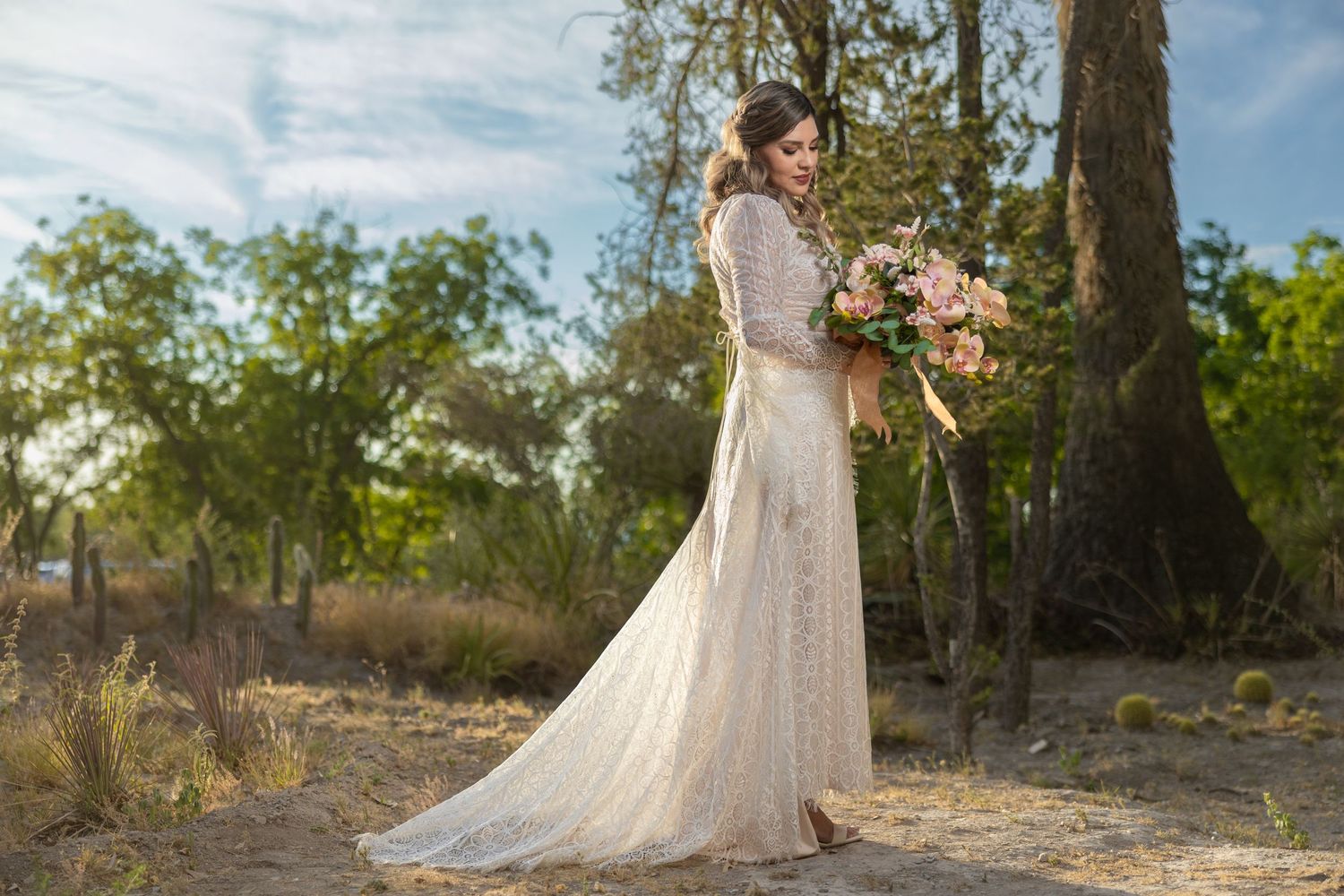 Full-length view of bride in flowing lace dress holding pink bouquet in woodland setting. boda en Parras Coahuila fotógrafo
